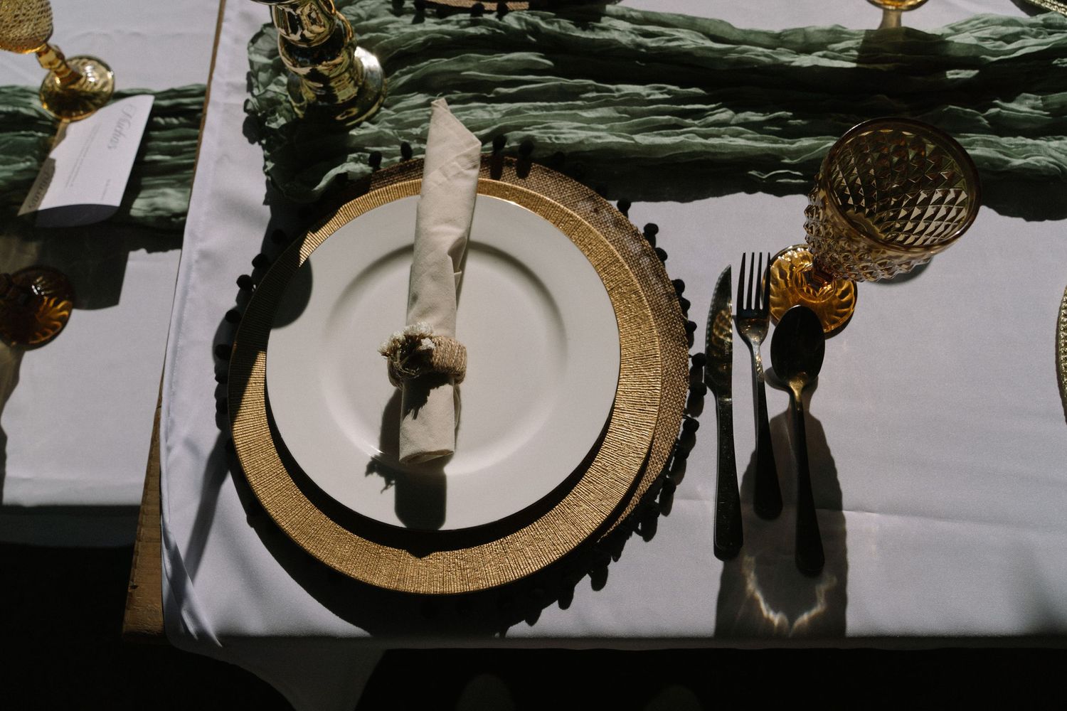Elegant gold-rimmed wedding place setting with white plate, folded napkin, amber glassware, and gold cutlery on white tablecloth.