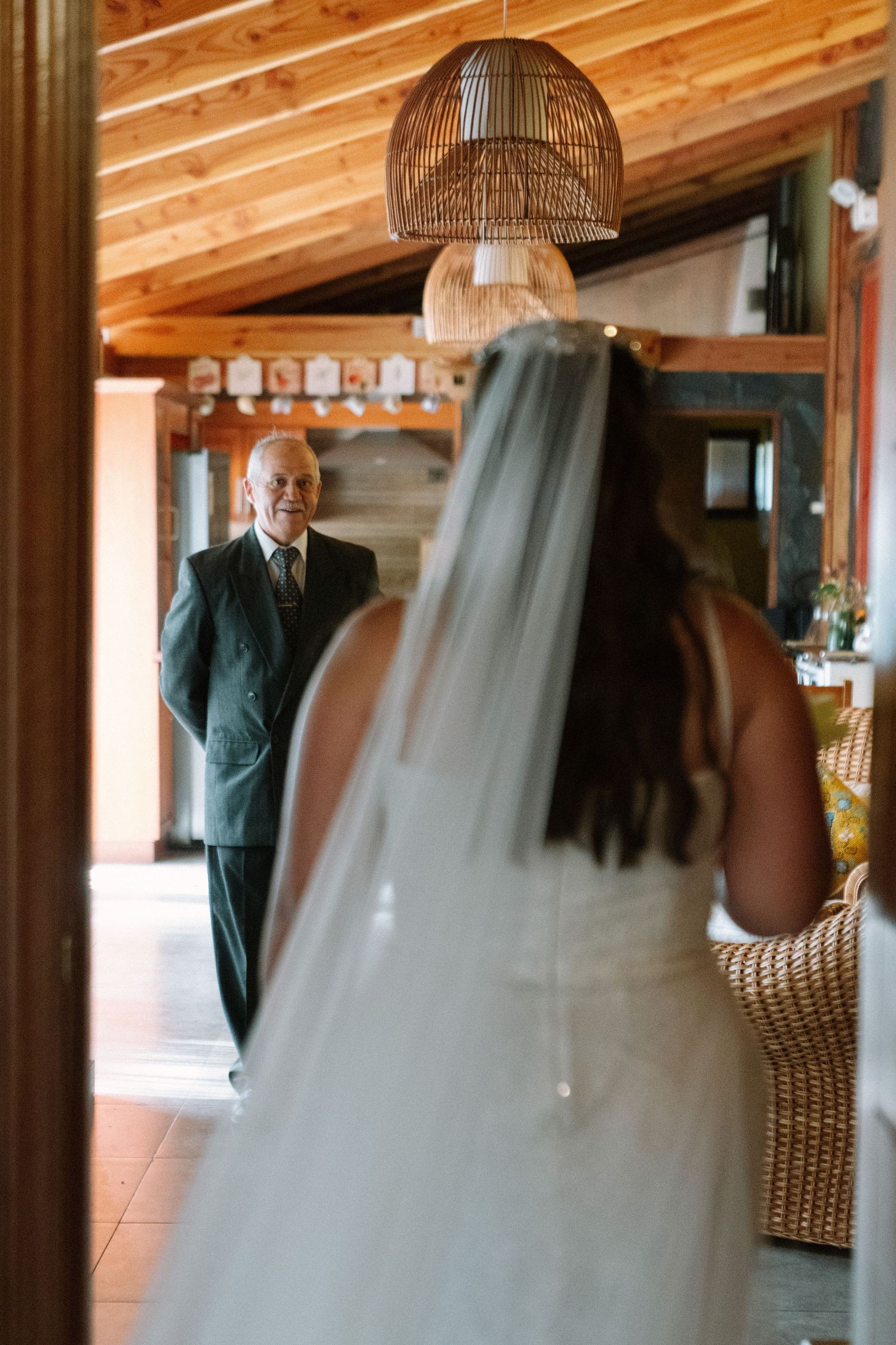 Bride in white gown and veil shares first look moment with older gentleman in suit inside rustic wooden venue.