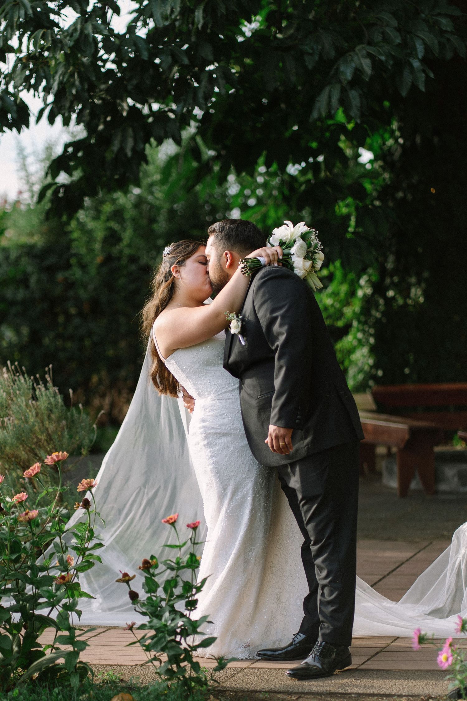Bride and groom share first kiss surrounded by roses and green foliage in an outdoor garden setting.