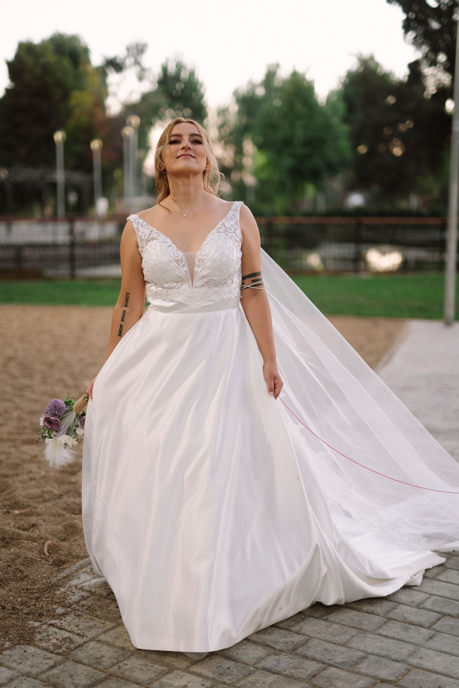 A radiant bride in a flowing white gown smiles while holding a purple bouquet outdoors.