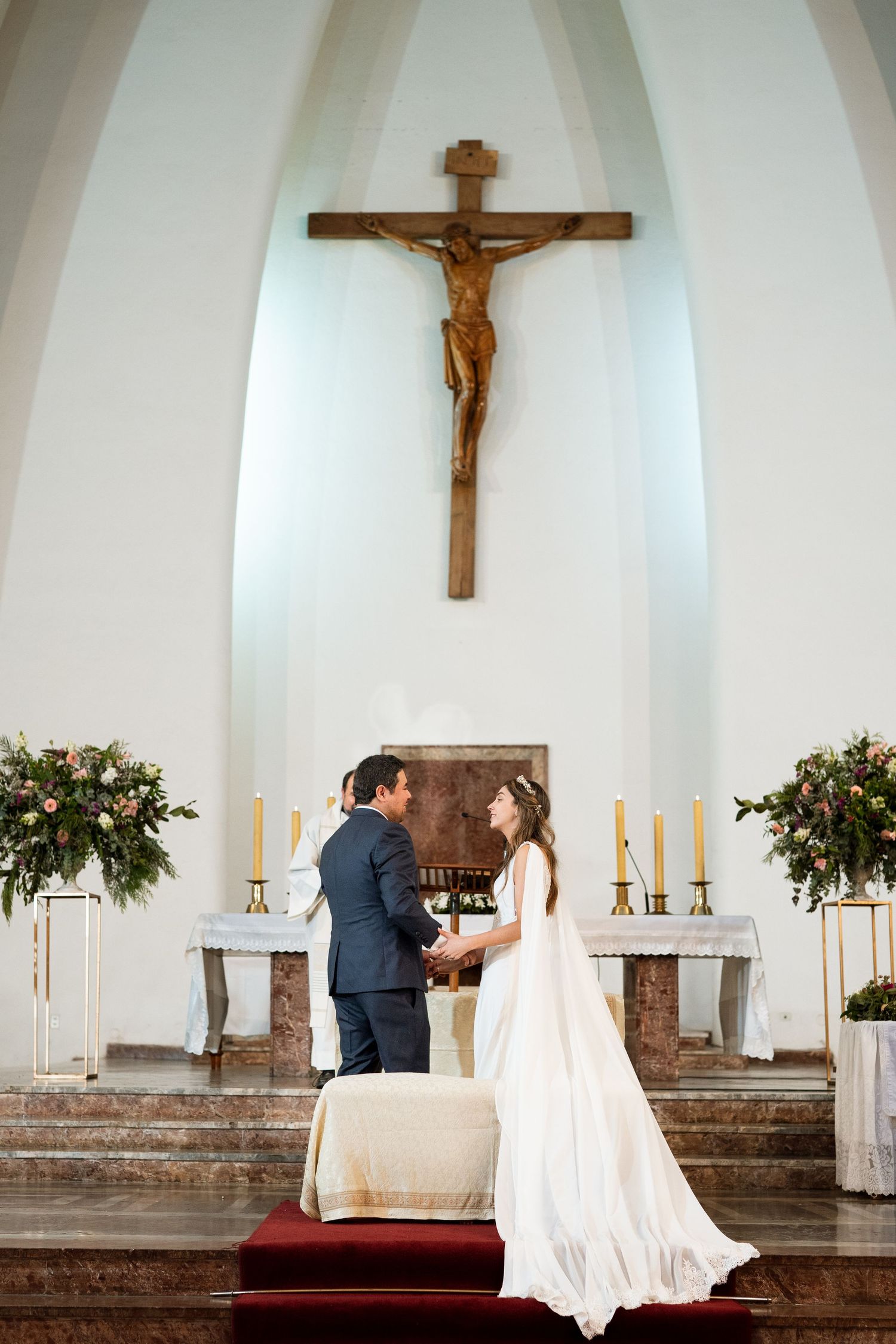 Wedding couple stands together on church altar steps with crucifix and floral arrangements.