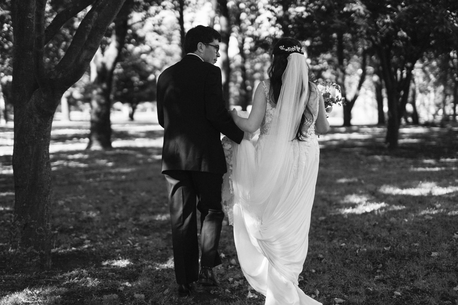 Black and white photo of newlyweds walking together through a tree-lined path.