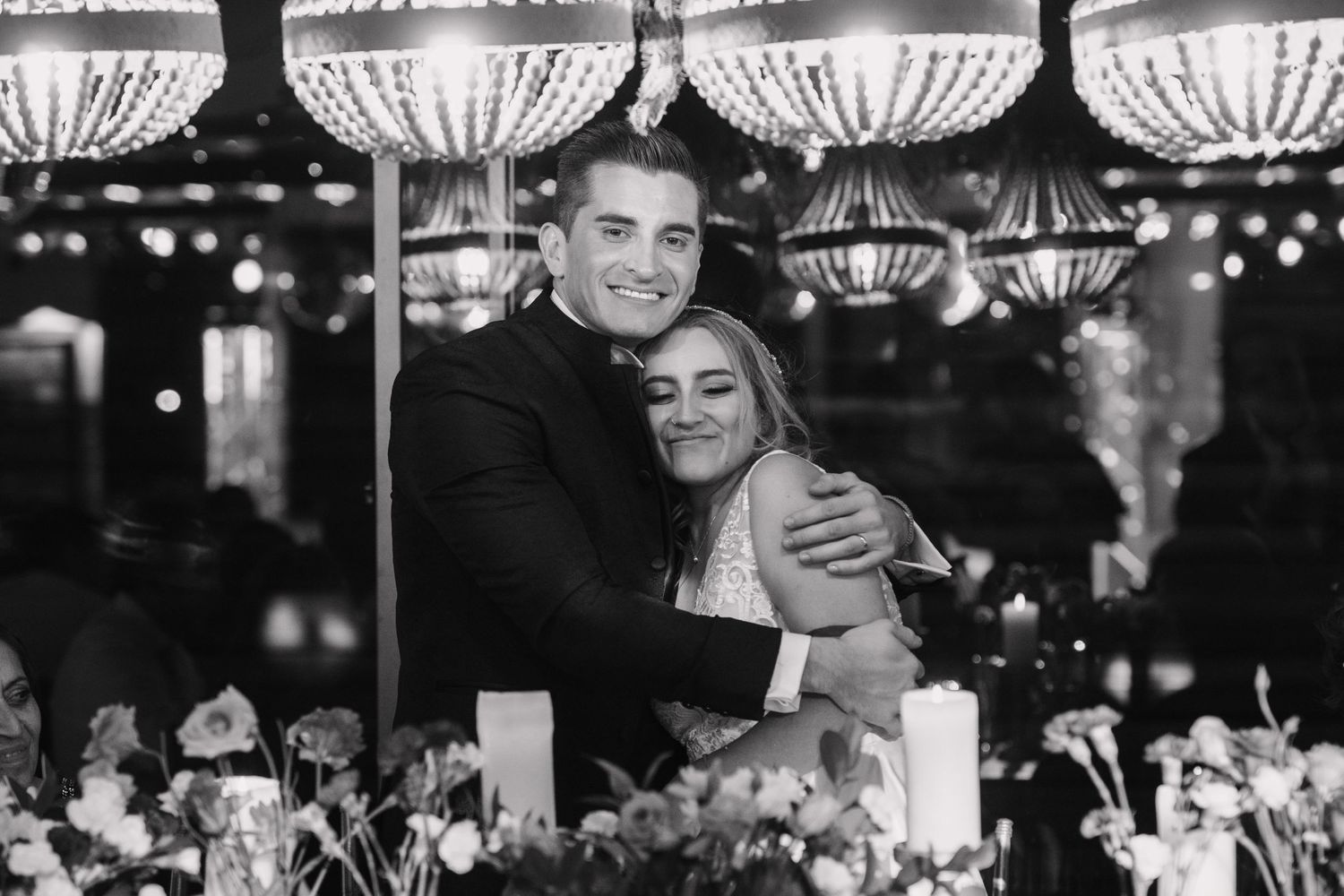 A romantic black and white photo of newlyweds embracing at their reception with crystal chandeliers overhead.