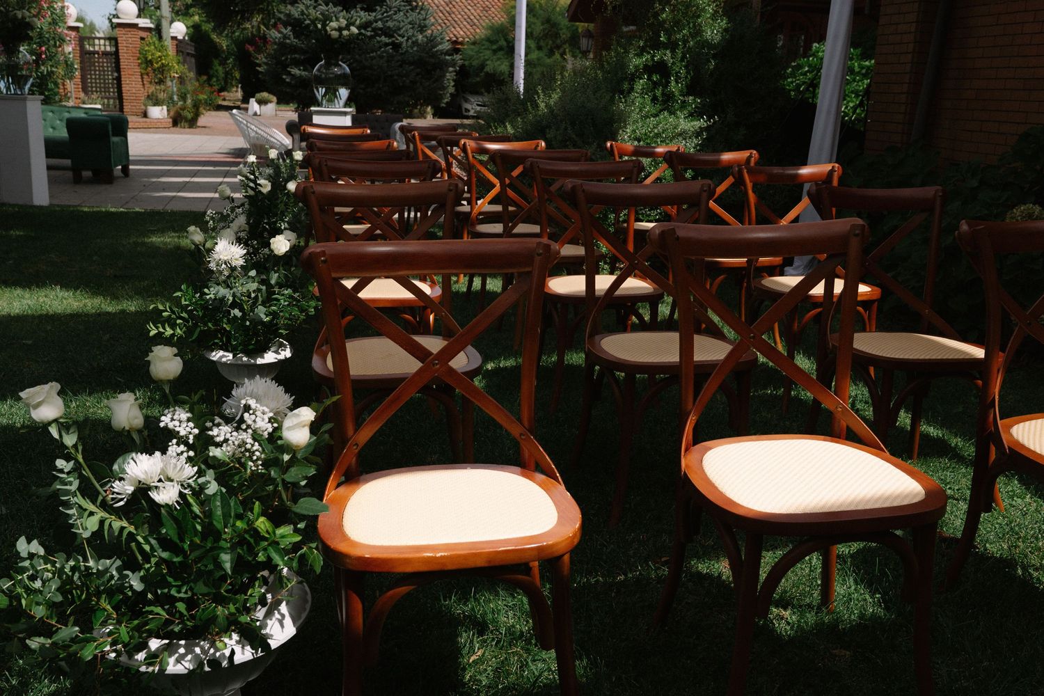 Wooden cross-back chairs with cream cushions arranged on green grass for an outdoor wedding ceremony.