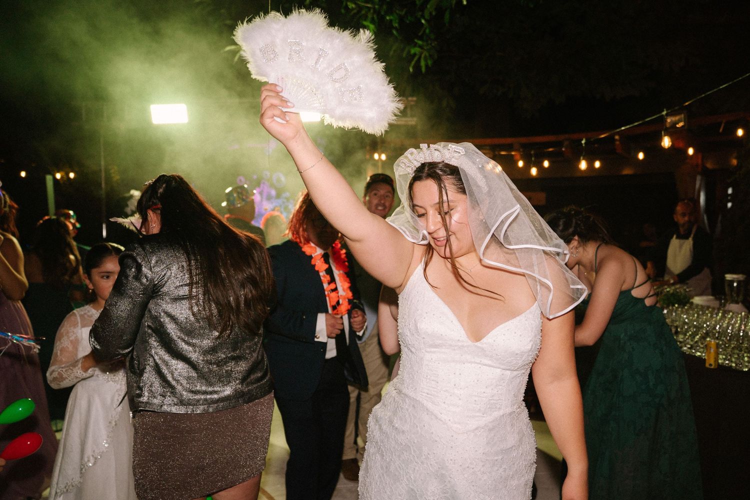 Bride in white wedding dress dancing joyfully with feather fan and veil at outdoor reception.