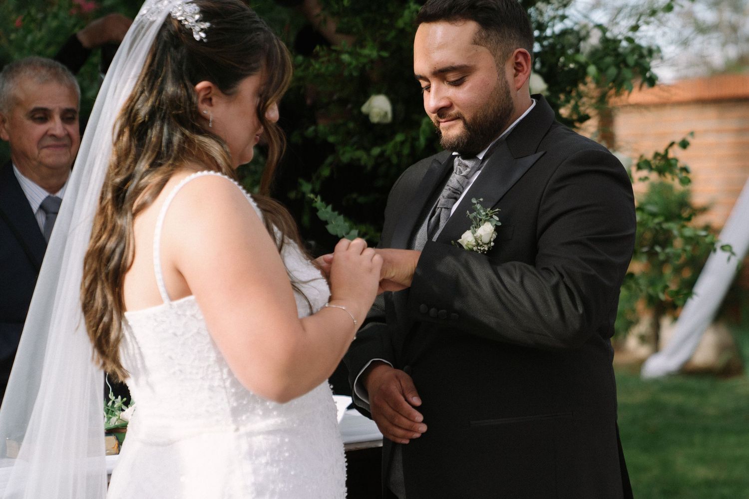 Groom placing wedding ring on brides finger during outdoor ceremony with guests watching.