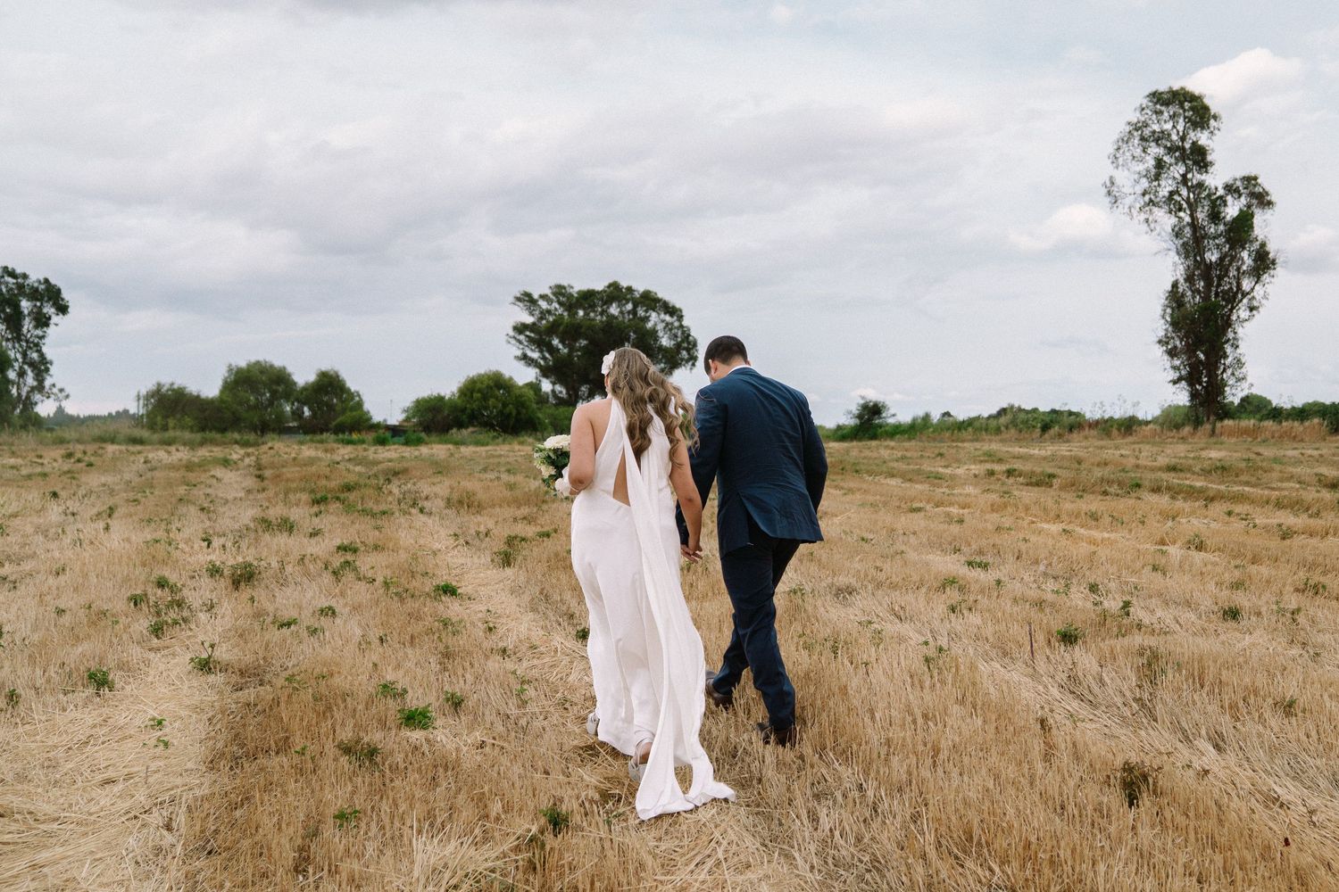 Bride and groom walking hand in hand through a golden field on their wedding day.
