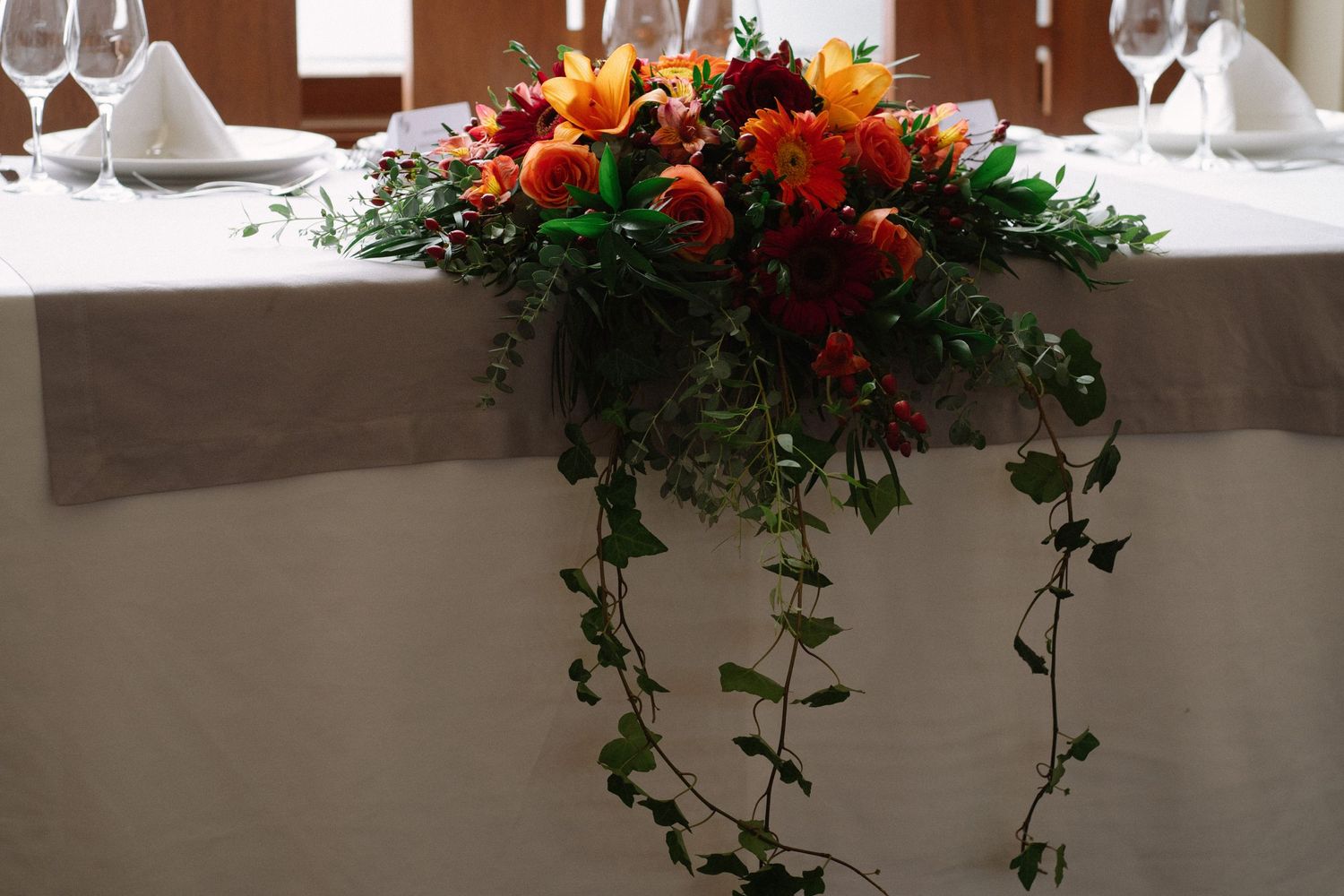 An elegant floral centerpiece with red and orange flowers cascades down a white tablecloth at a formal place setting.