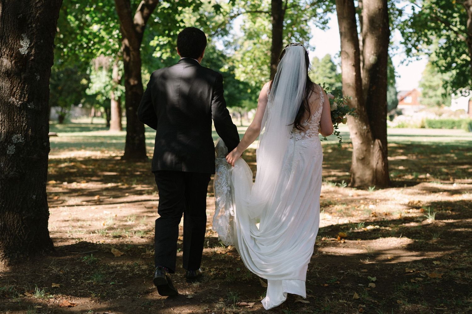 Couple walking hand in hand through a sunlit park on their wedding day.