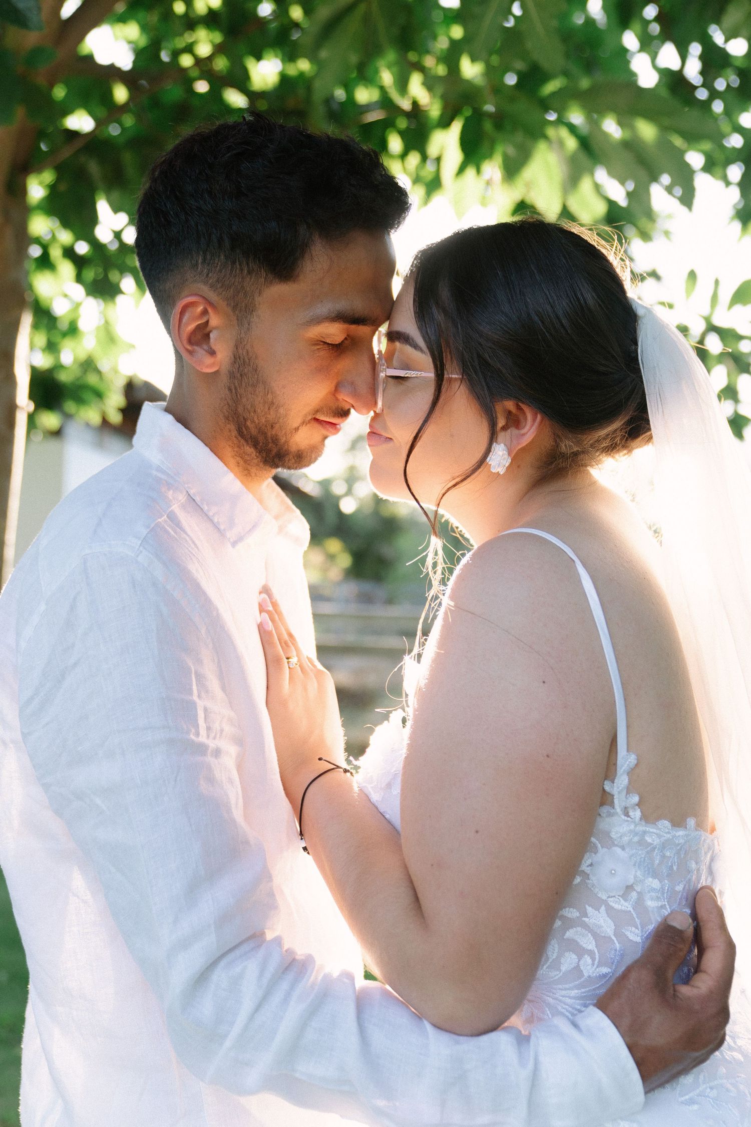 A bride and groom share an intimate forehead-to-forehead moment in a sun-drenched garden setting.