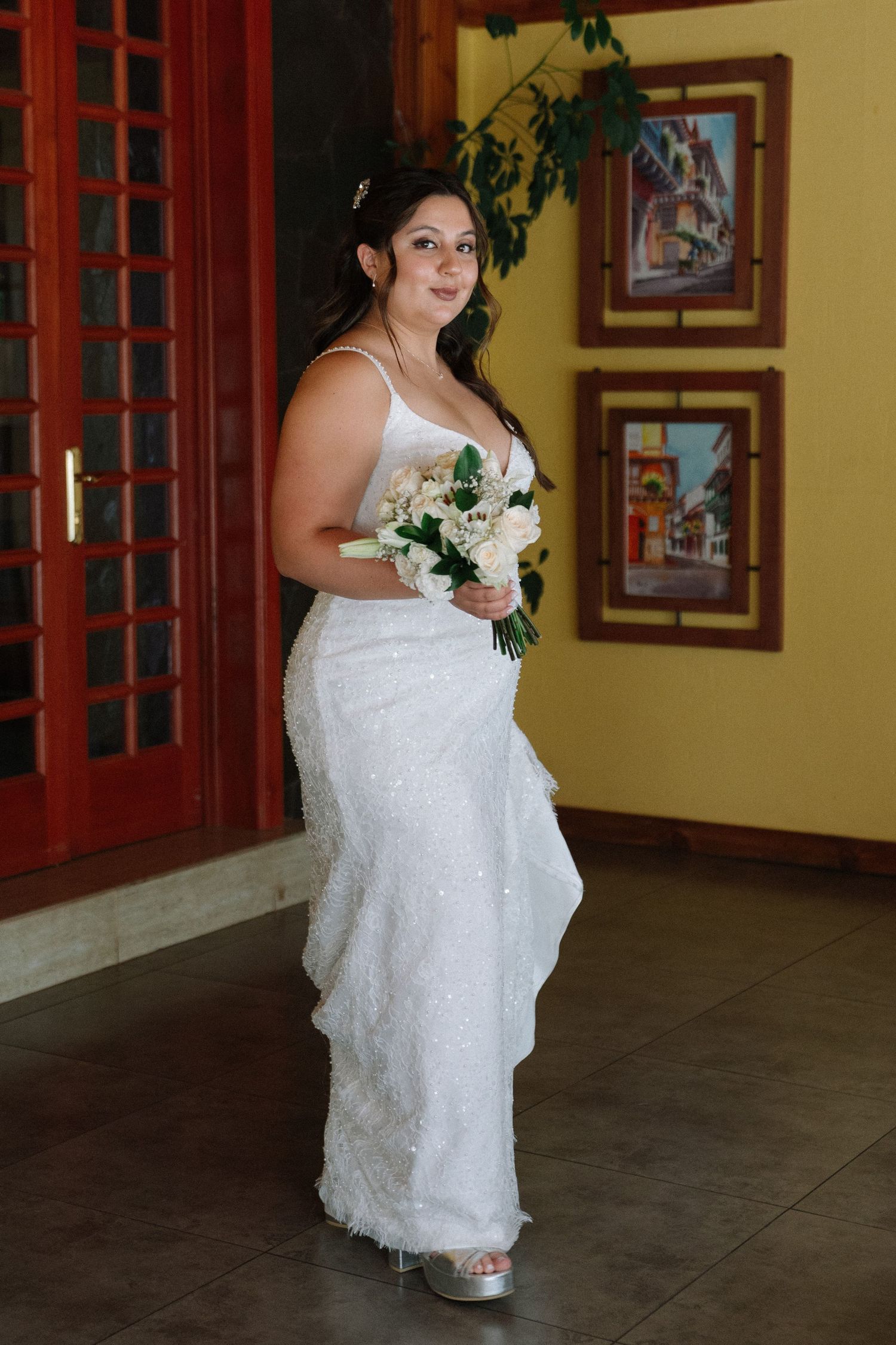 Bride in white beaded gown holding bouquet of white roses standing indoors before wedding ceremony.