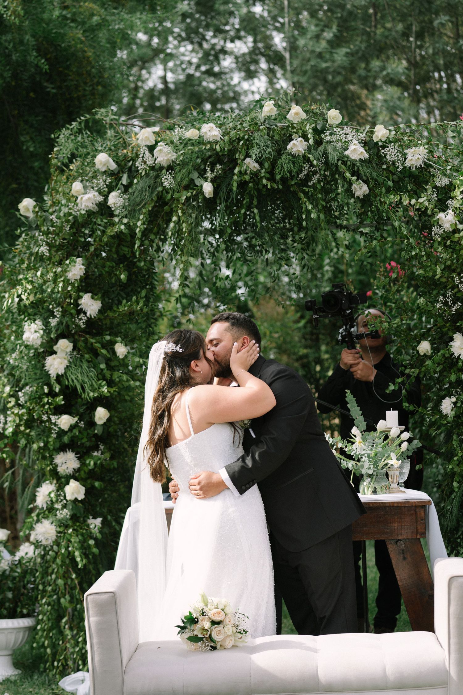 Bride and groom sharing their first kiss under a lush greenery and white floral arch.