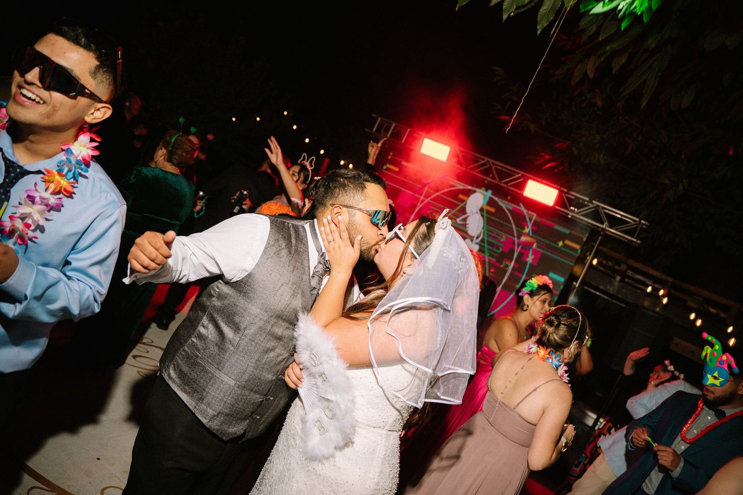 Bride and groom kissing on the dance floor surrounded by guests at a nighttime reception.