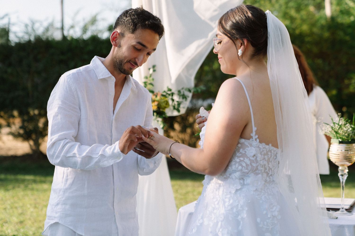 A groom in white places a ring on his bride's finger during an outdoor garden wedding ceremony.