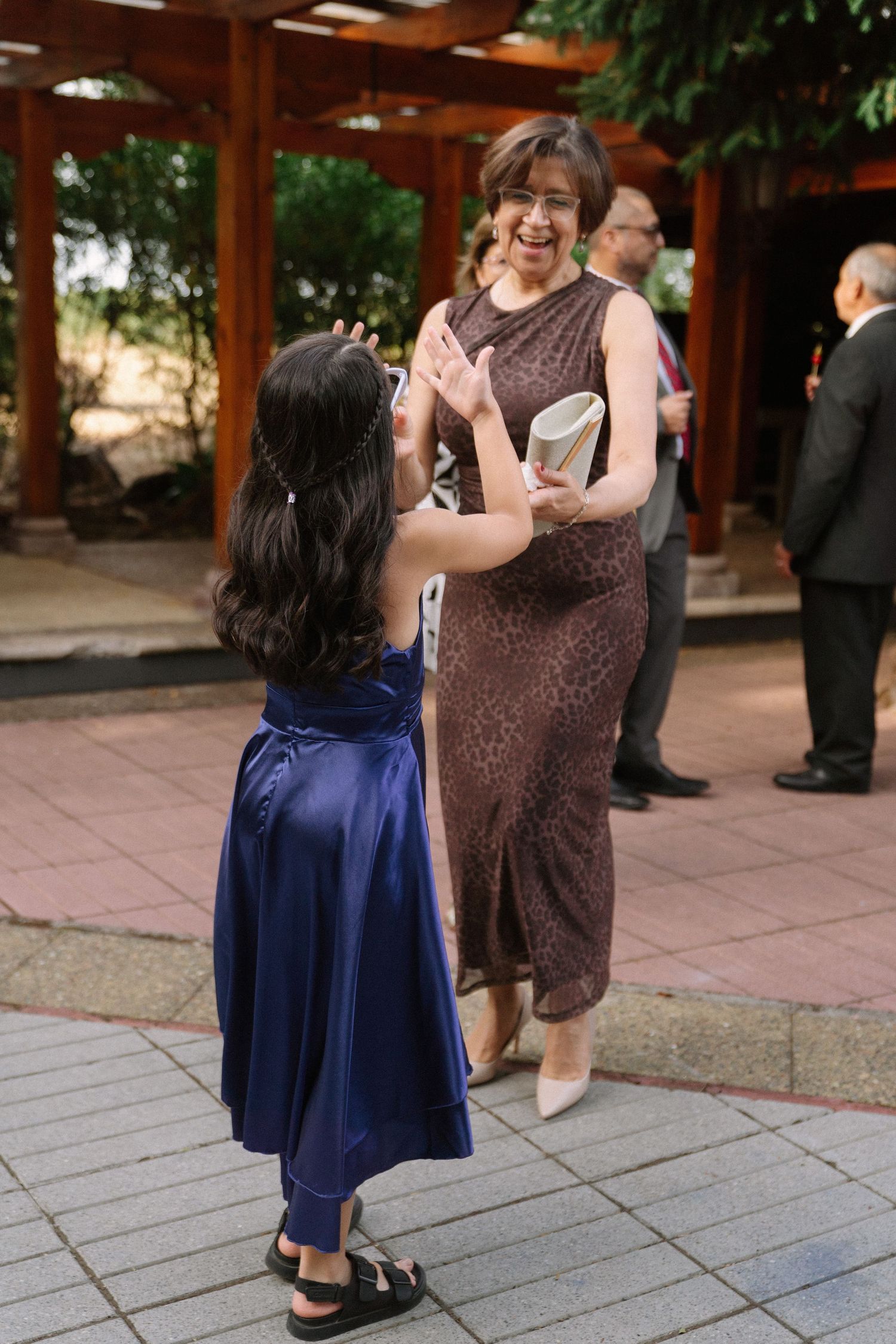 A smiling grandmother in a brown dress plays patty-cake with a young flower girl in a blue dress.