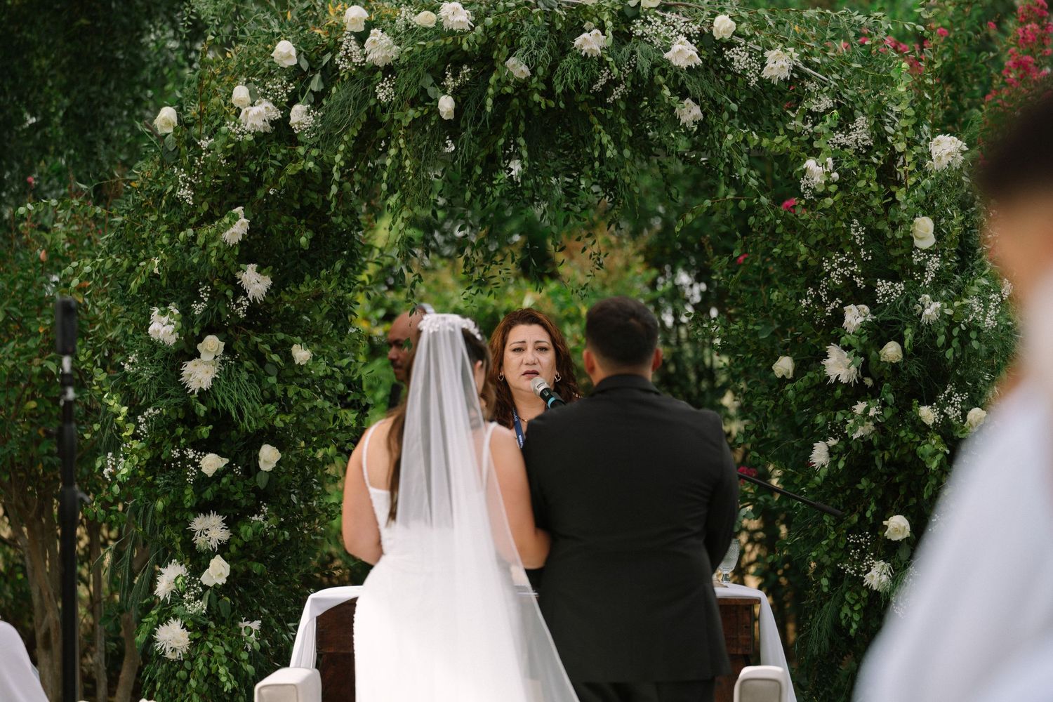 Bride and groom exchange vows beneath a lush green arch adorned with white roses at an outdoor wedding ceremony.
