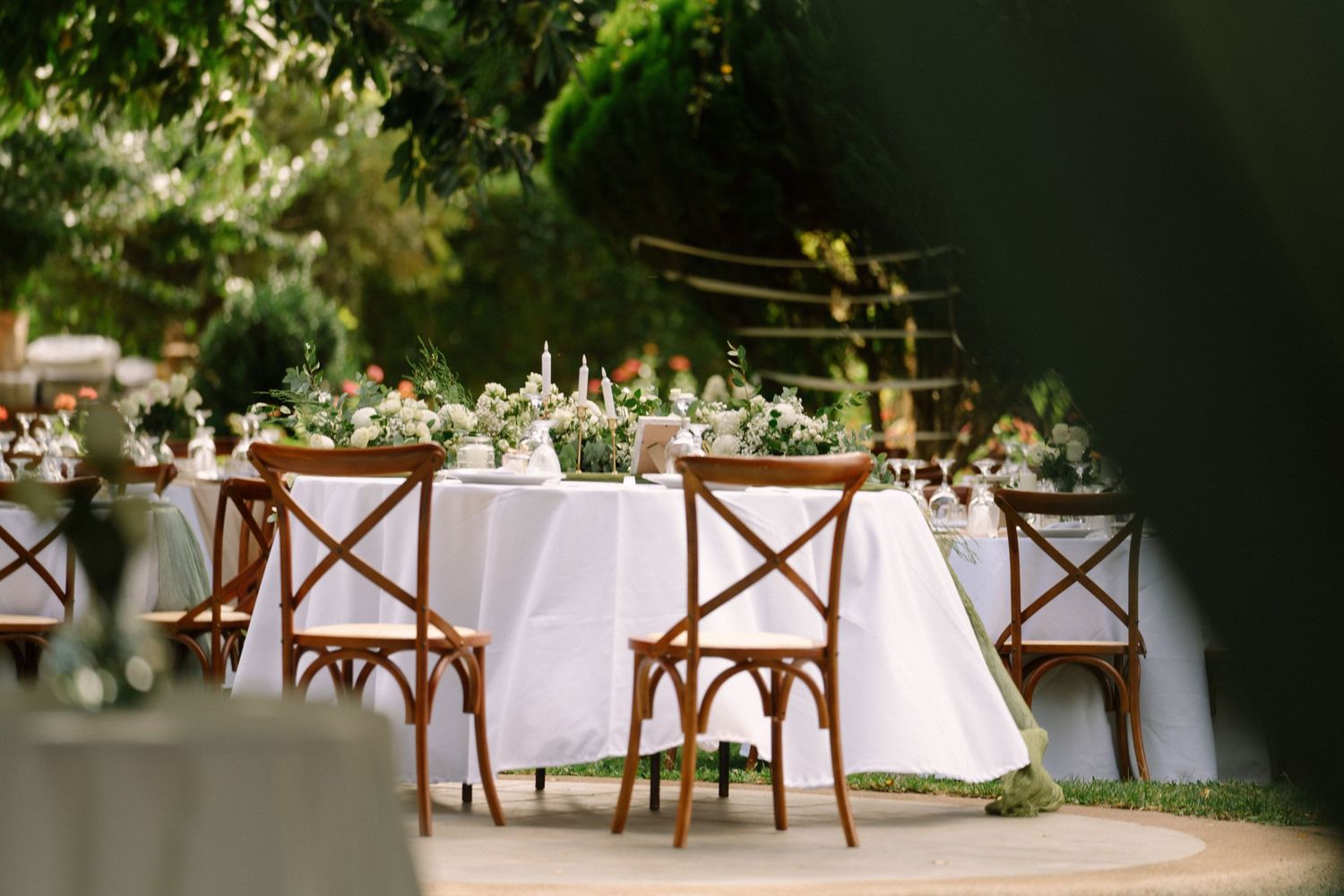 Elegant outdoor wedding reception table with white linens, greenery centerpieces, and rustic wooden cross-back chairs in a garden.