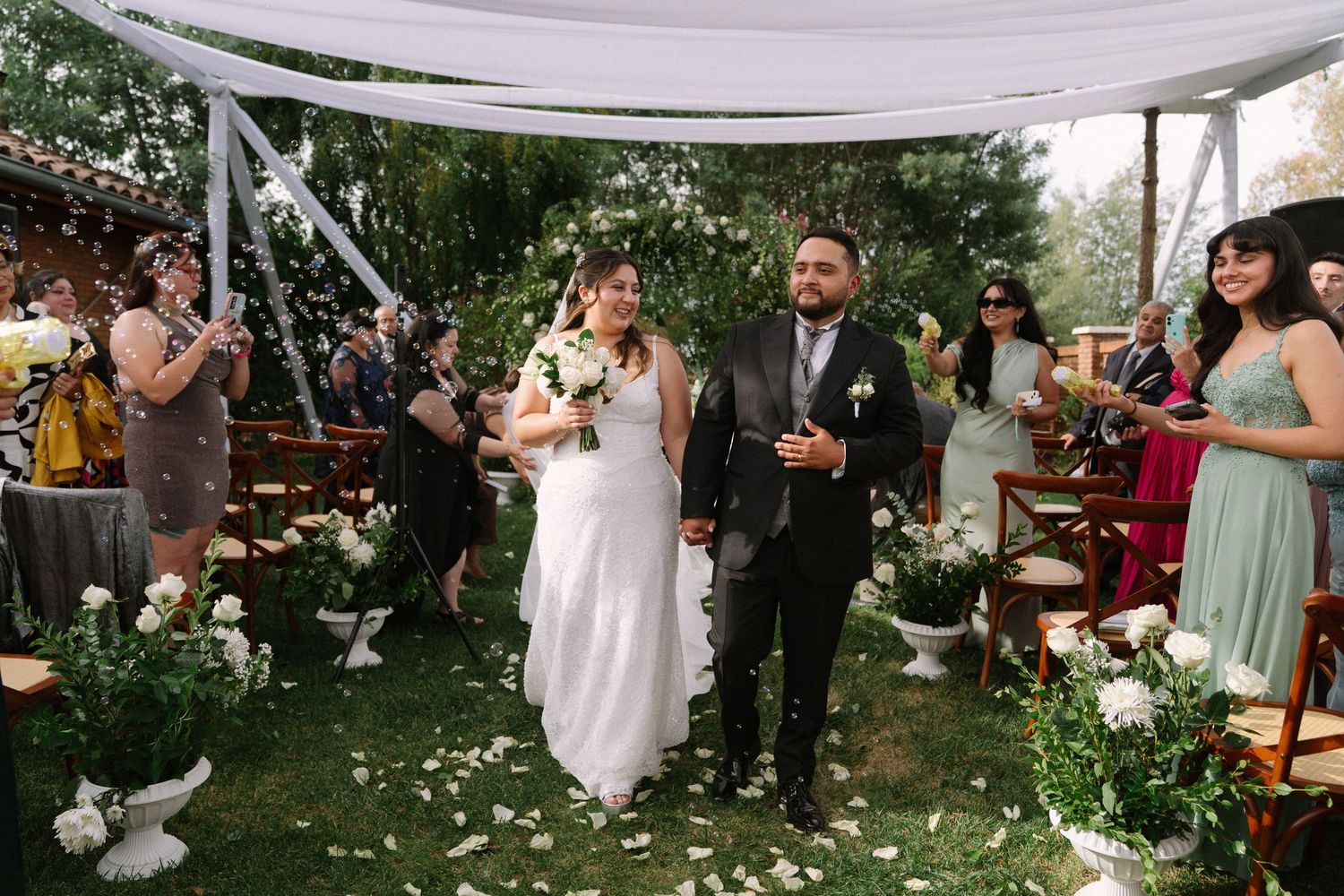 Bride and groom walk joyfully down the petal-lined aisle as guests celebrate at an outdoor garden wedding.