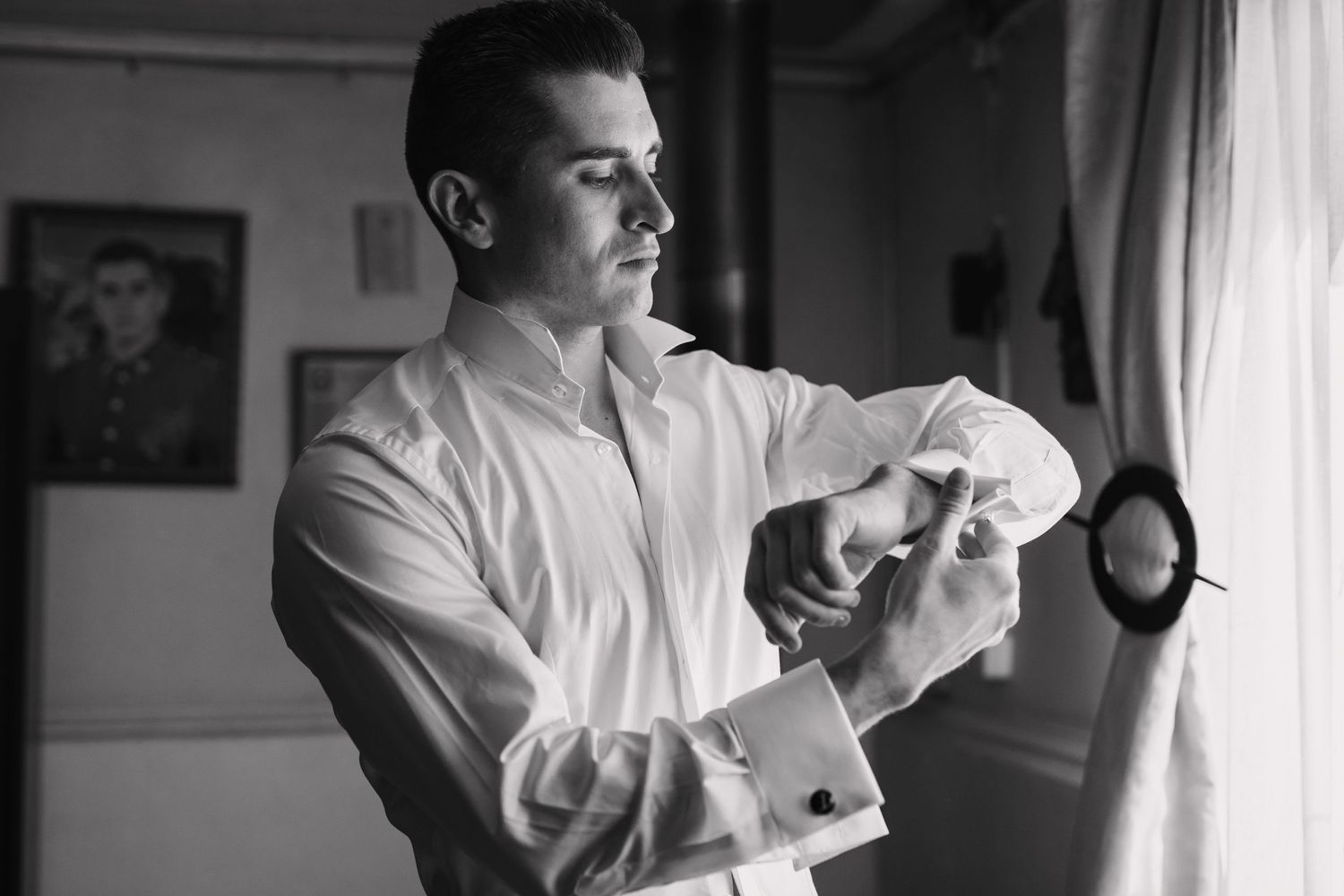 Person wearing white dress shirt adjusting their sleeve cuff in a room by a window.