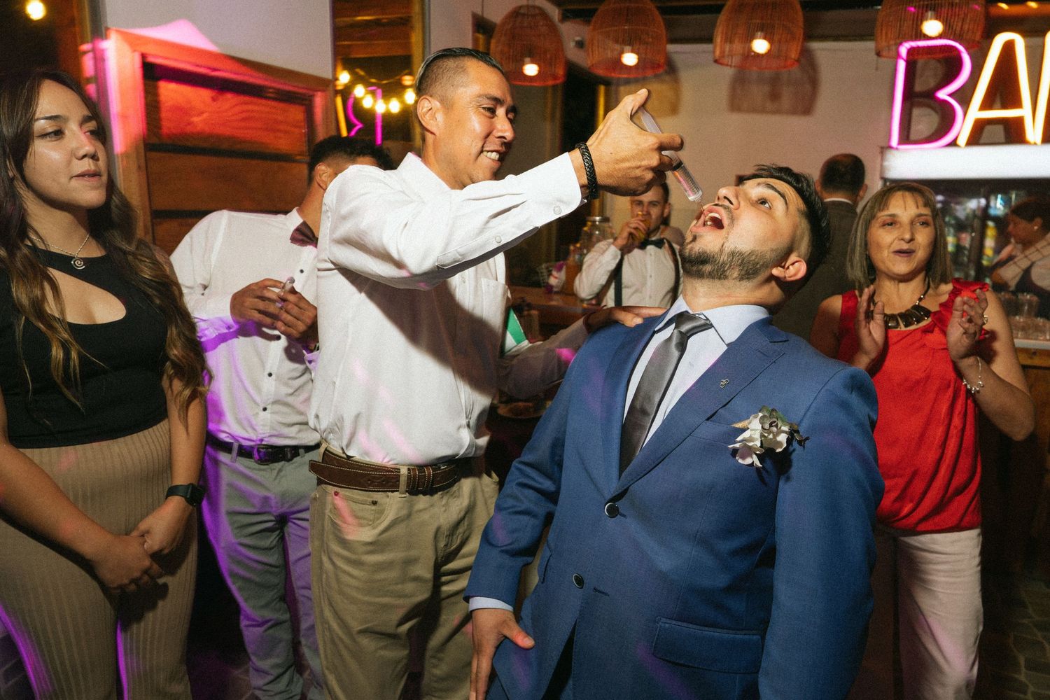 A man pours a drink into another's mouth at a lively bar celebration.