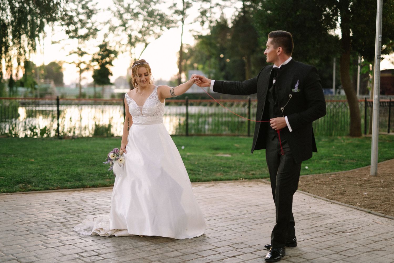 Wedding couple dances playfully on a paved pathway in a park setting at sunset.