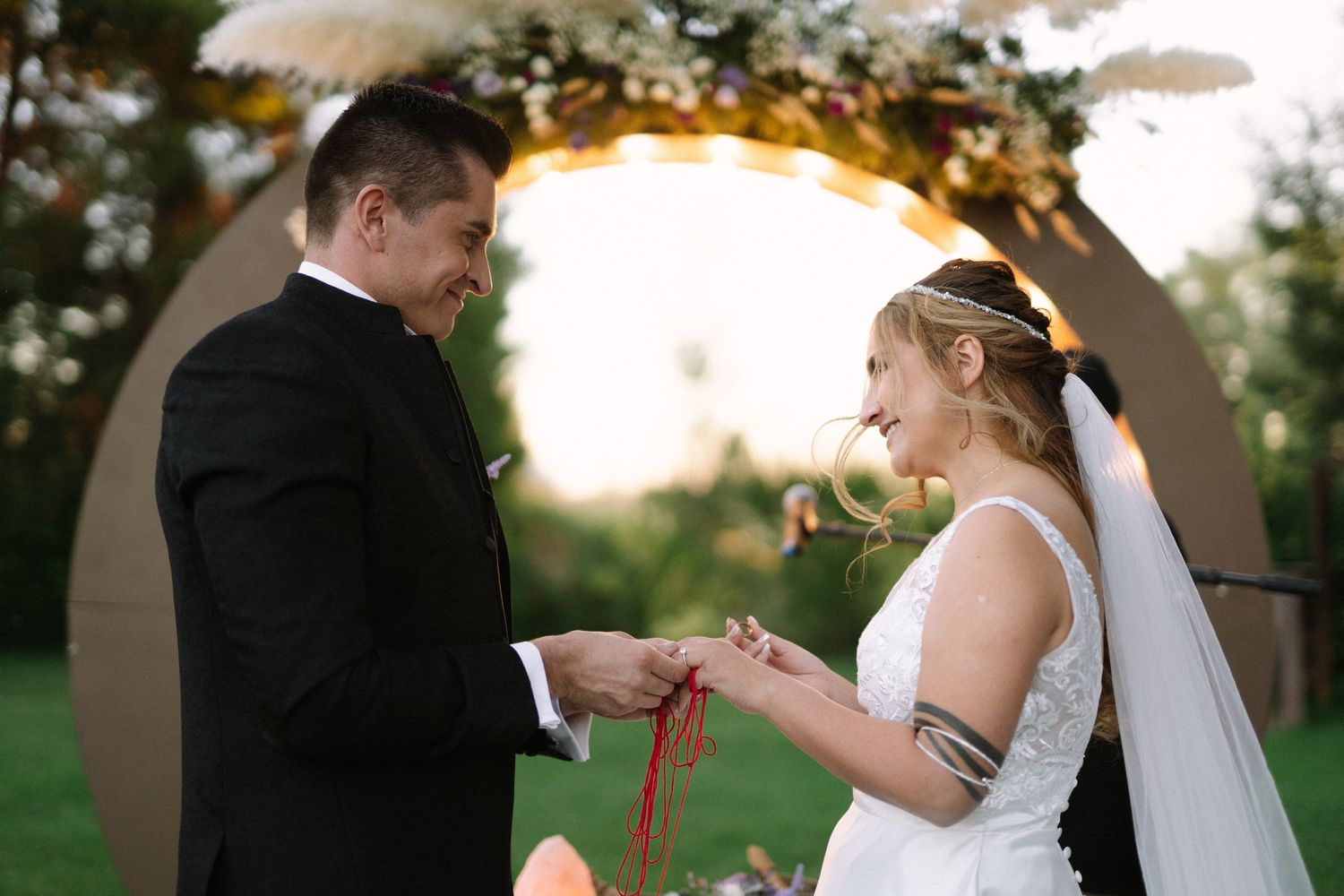 Newlyweds share a joyful moment during ring exchange beneath a decorated ceremony arch at sunset.