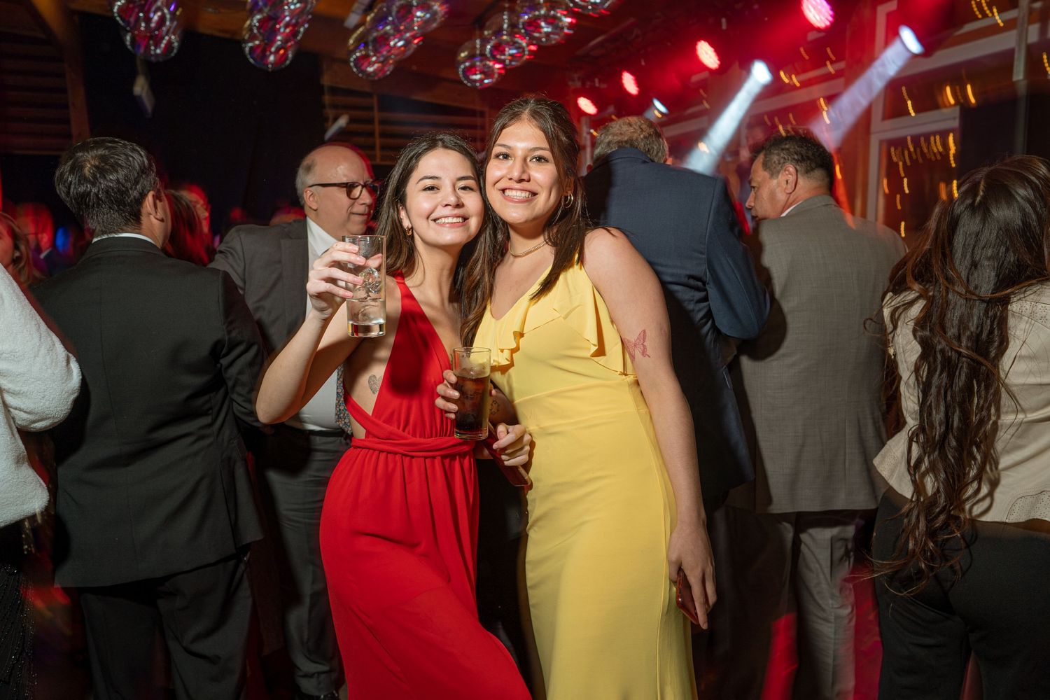 Two women in formal dresses, one red and one yellow, enjoying drinks at an evening celebration.