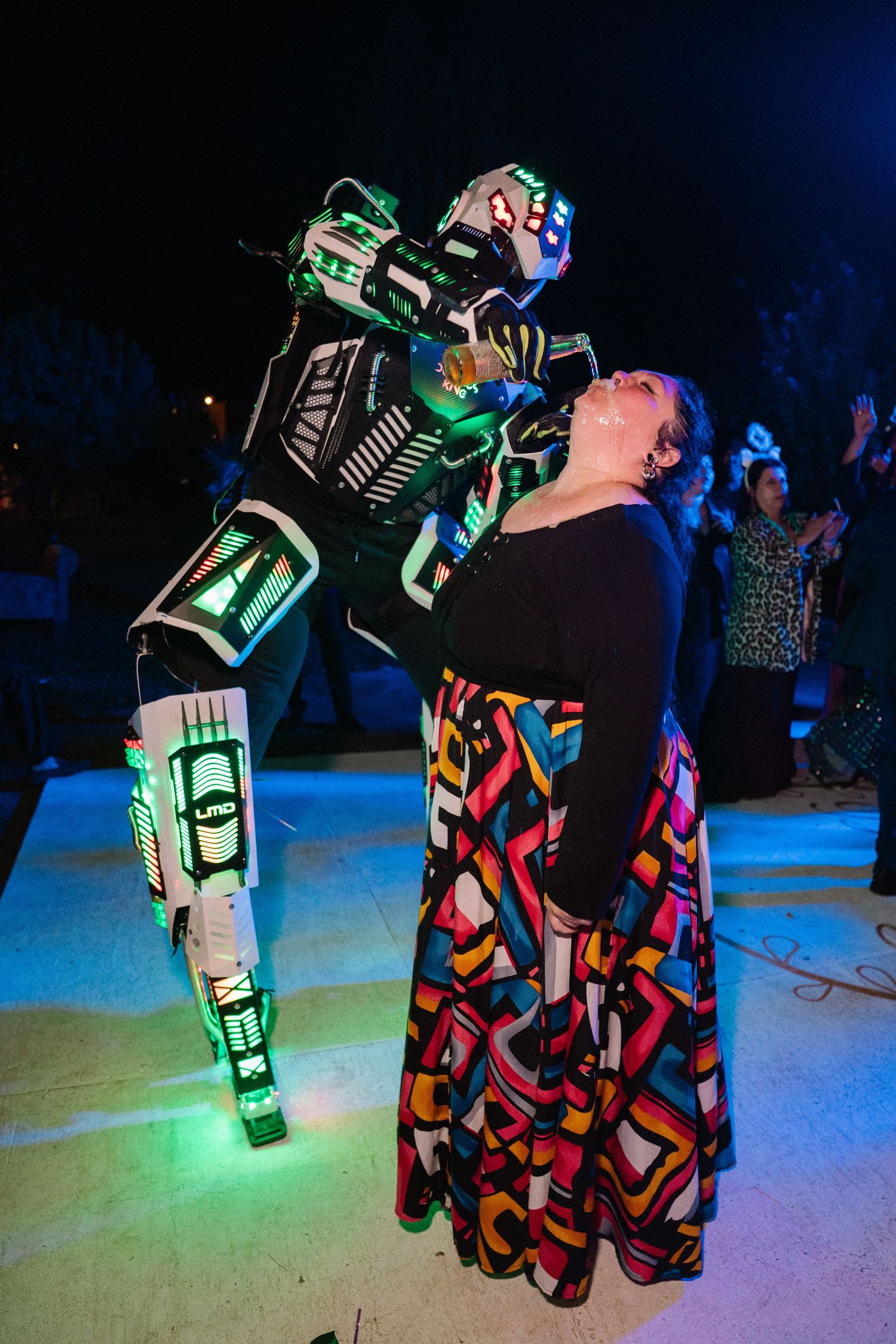 Woman in colorful dress poses with illuminated LED robot costume performer at nighttime event.