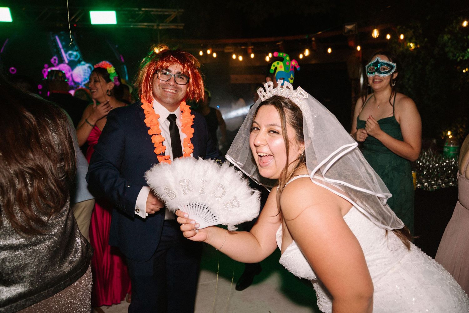 Bride laughing with fan while guest wears colorful costume at festive wedding reception.