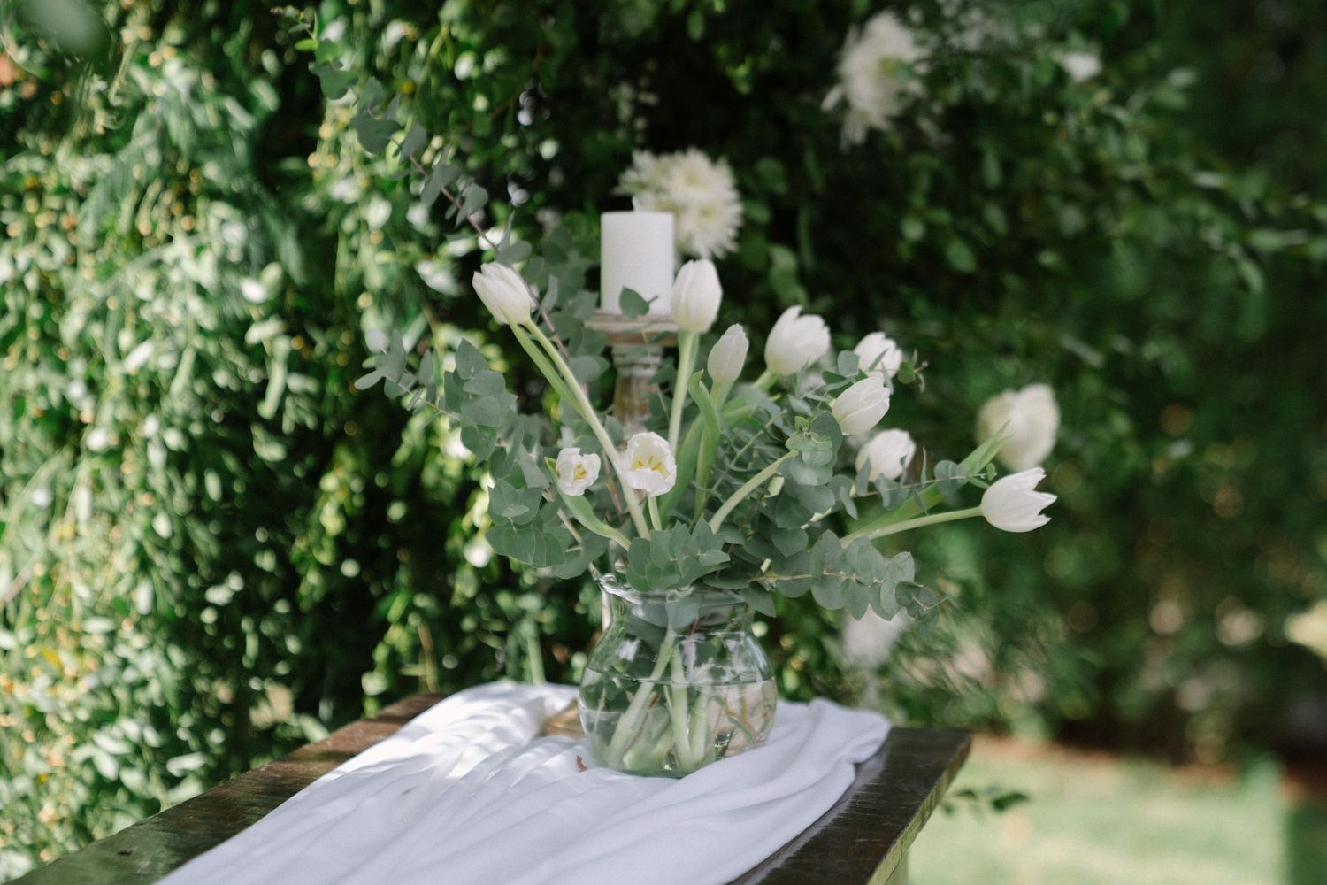 White tulips and greenery in a glass vase with a candle centerpiece on a draped table at an outdoor wedding.