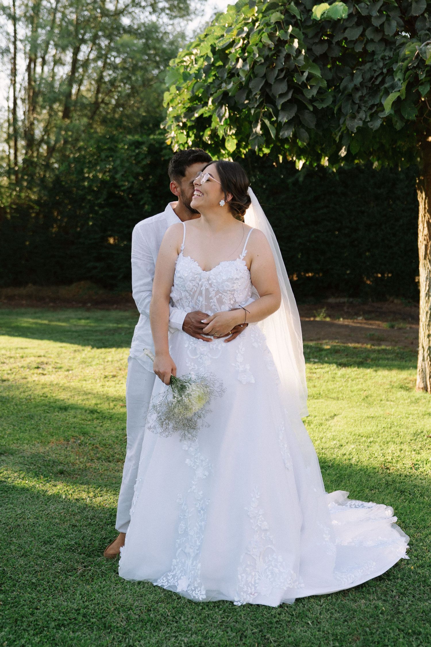 A groom embraces his smiling bride in a lace wedding gown holding a bouquet in a sunny garden.