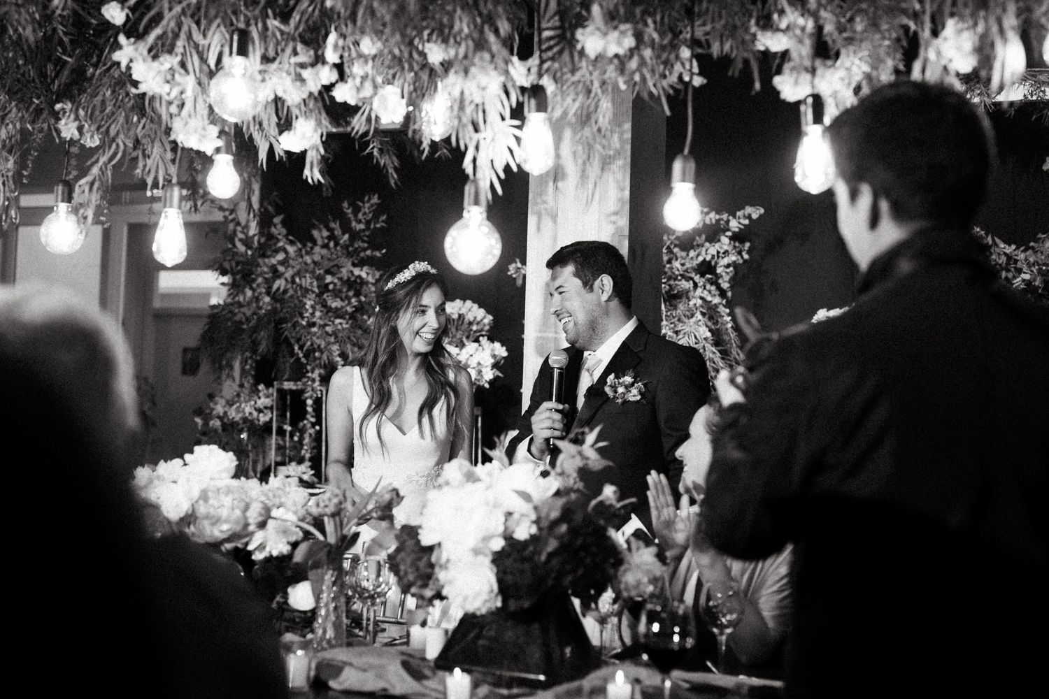 Black and white photo of wedding reception with hanging lights and floral decor.