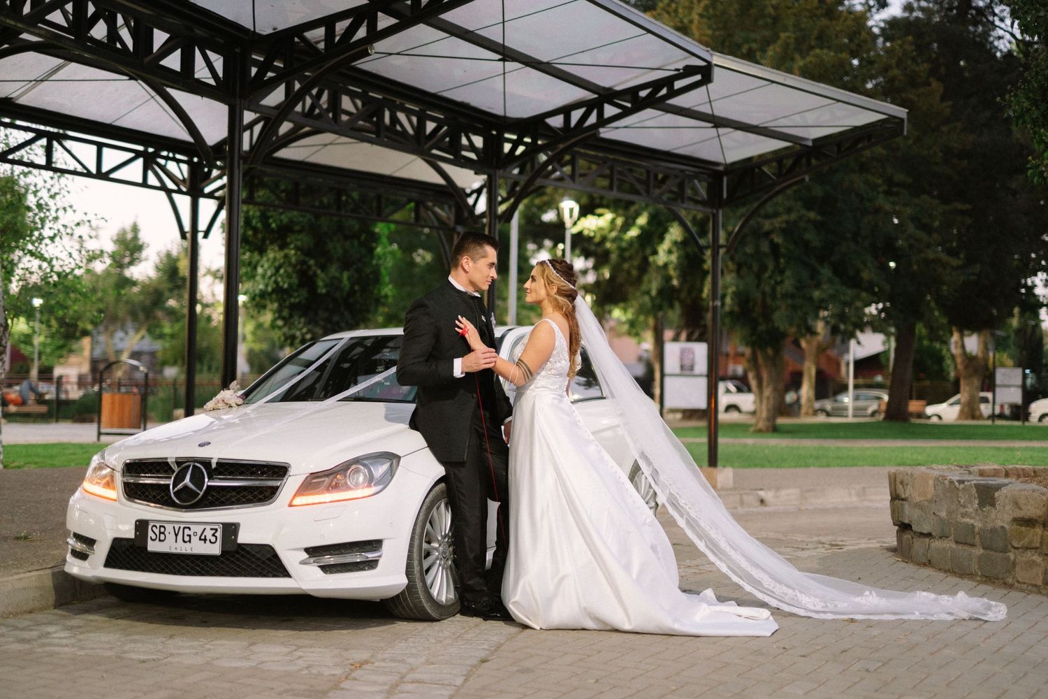 A newlywed couple poses with their white Mercedes-Benz under a covered pavilion.