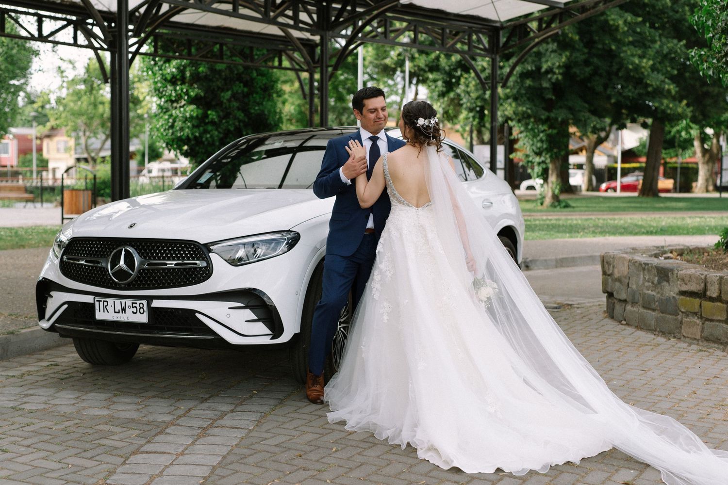 Wedding couple poses beside silver Mercedes-Benz car in formal attire.