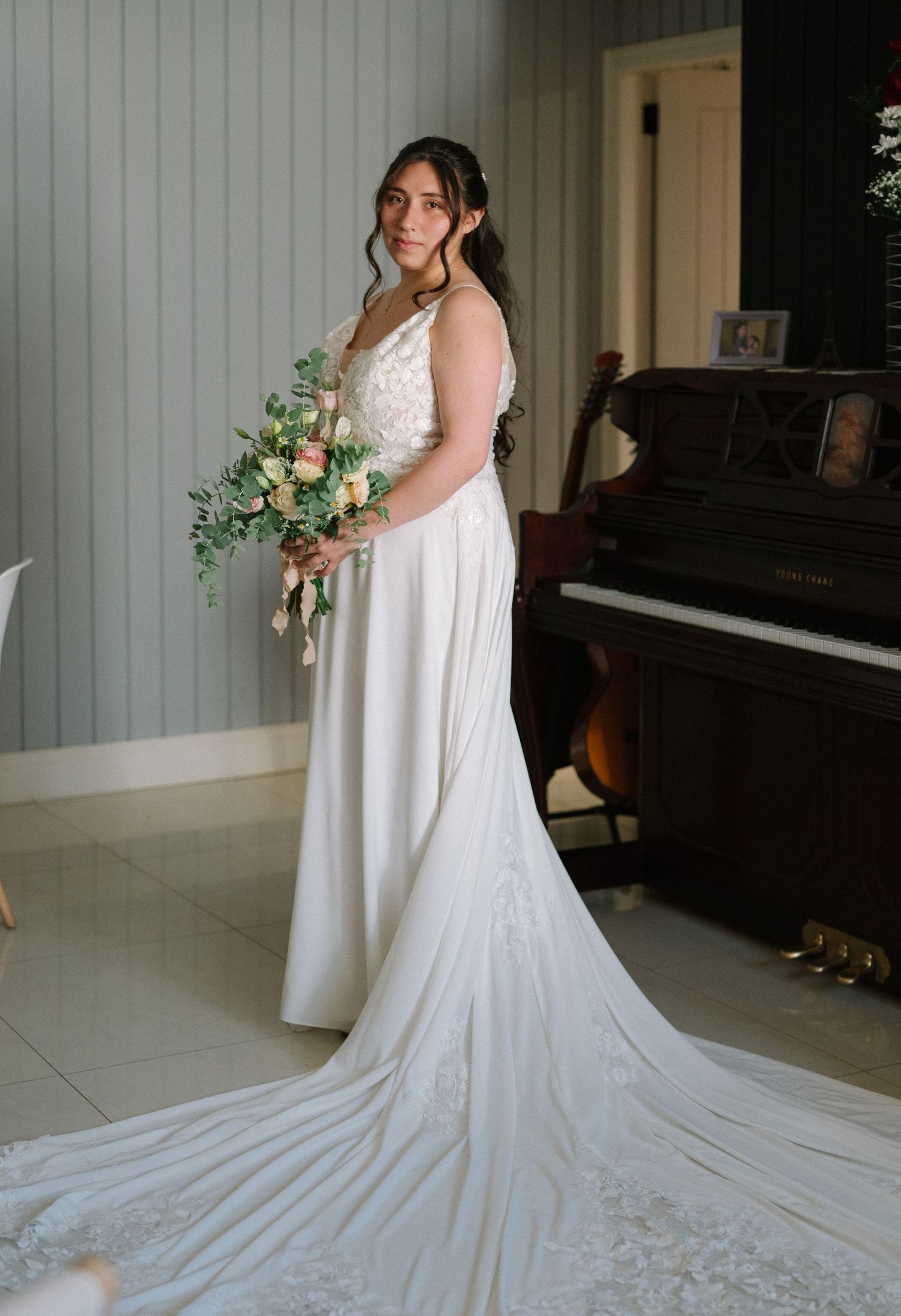 Bride in flowing white gown with train holds elegant bouquet beside piano.