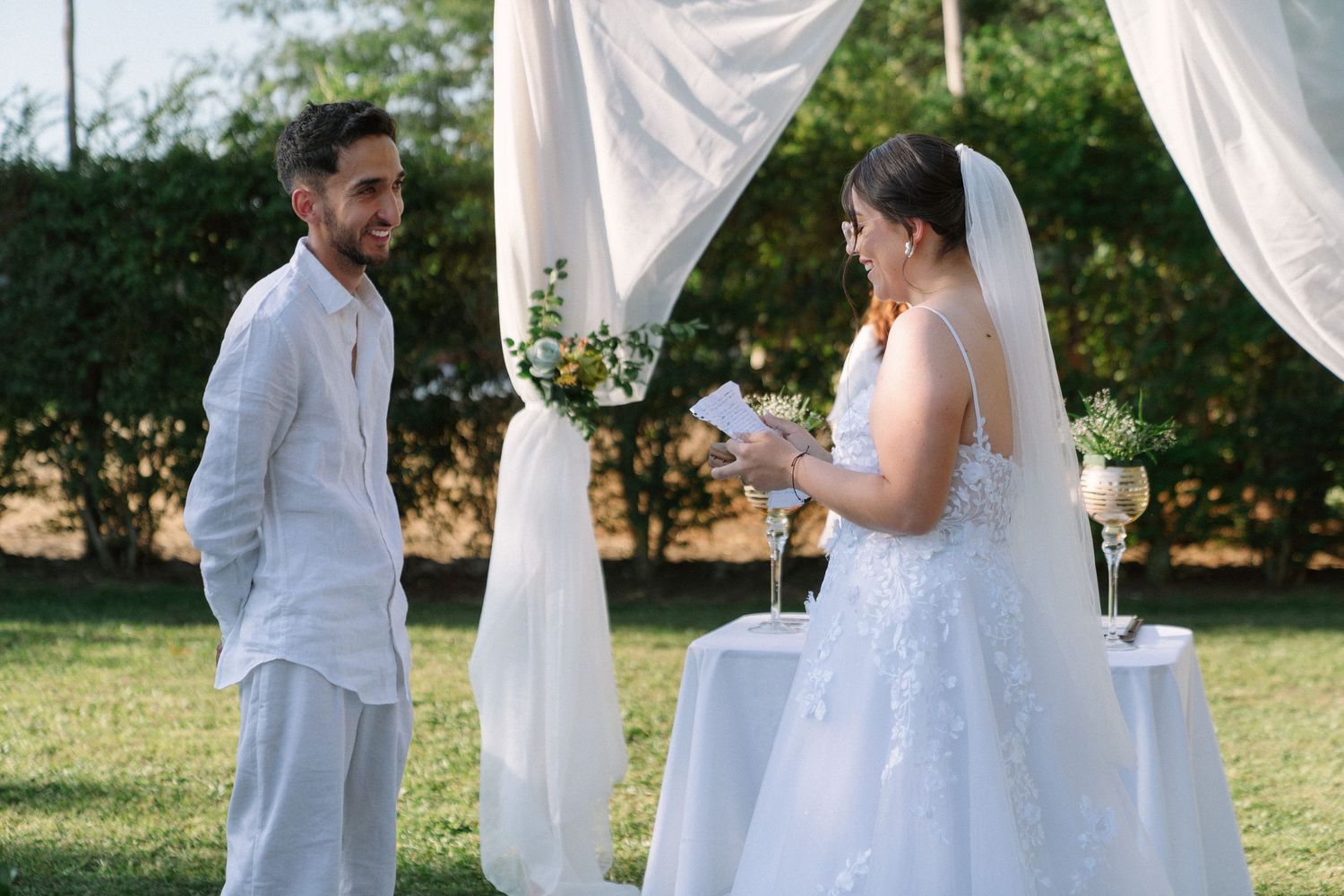 Bride reads vows to groom at outdoor wedding ceremony with white drape arch and floral arrangements.