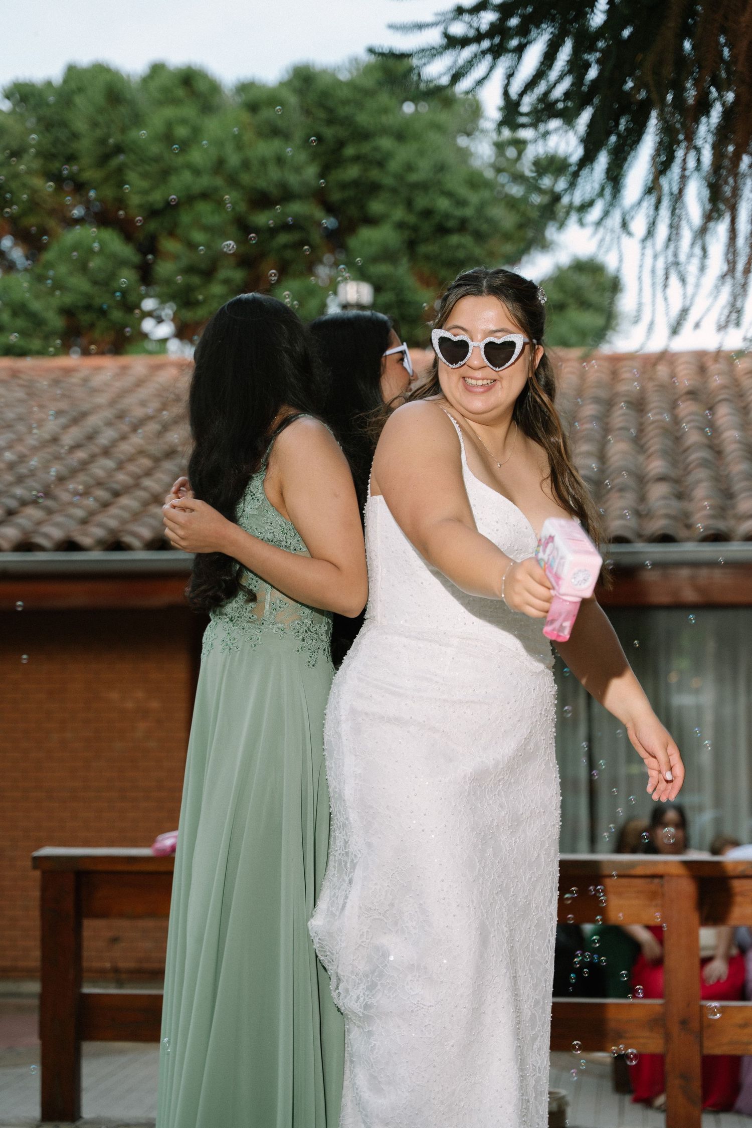 Smiling bride in white dress and heart sunglasses poses with bridesmaid in green gown at wedding.