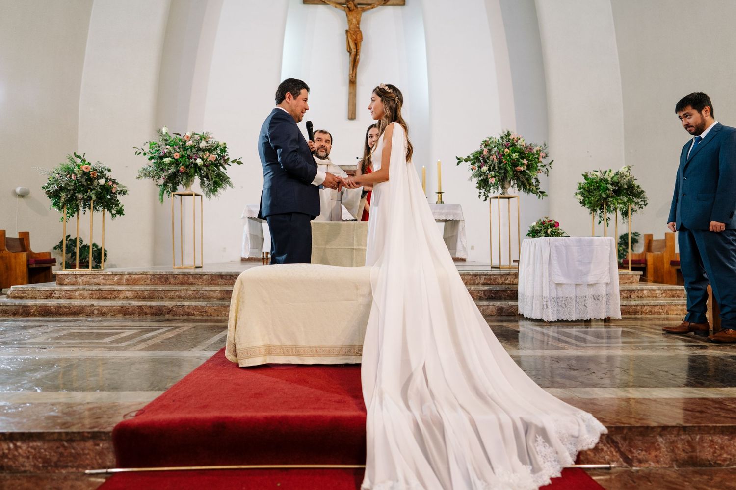 A couple holds hands during their wedding ceremony in front of a crucifix and floral arrangements.