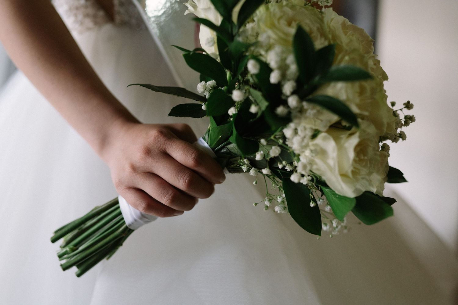 Close-up of hands holding elegant white and green wedding bouquet with ribbon wrap.
