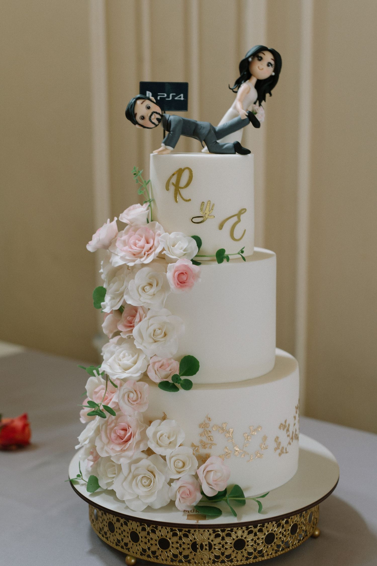 White wedding cake decorated with pink roses and playful cake topper on gold stand.