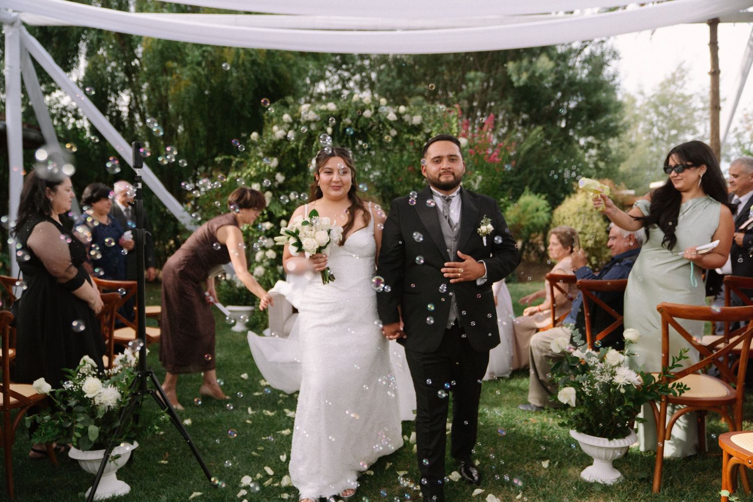 Newlyweds walk down the aisle as guests toss flower petals and bubbles at their outdoor garden wedding ceremony.