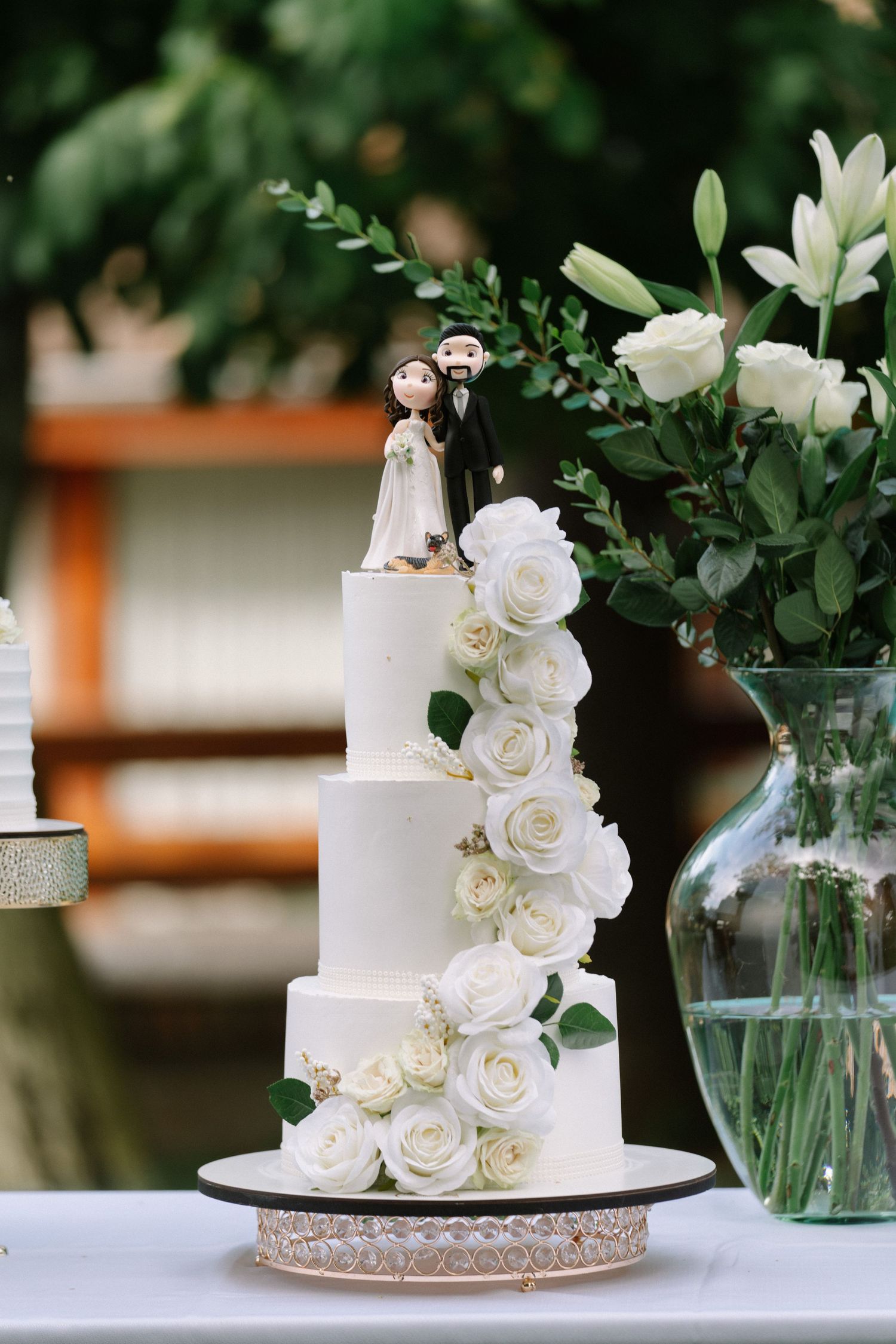 Elegant three-tier white wedding cake with roses and bride and groom cake toppers outdoors.
