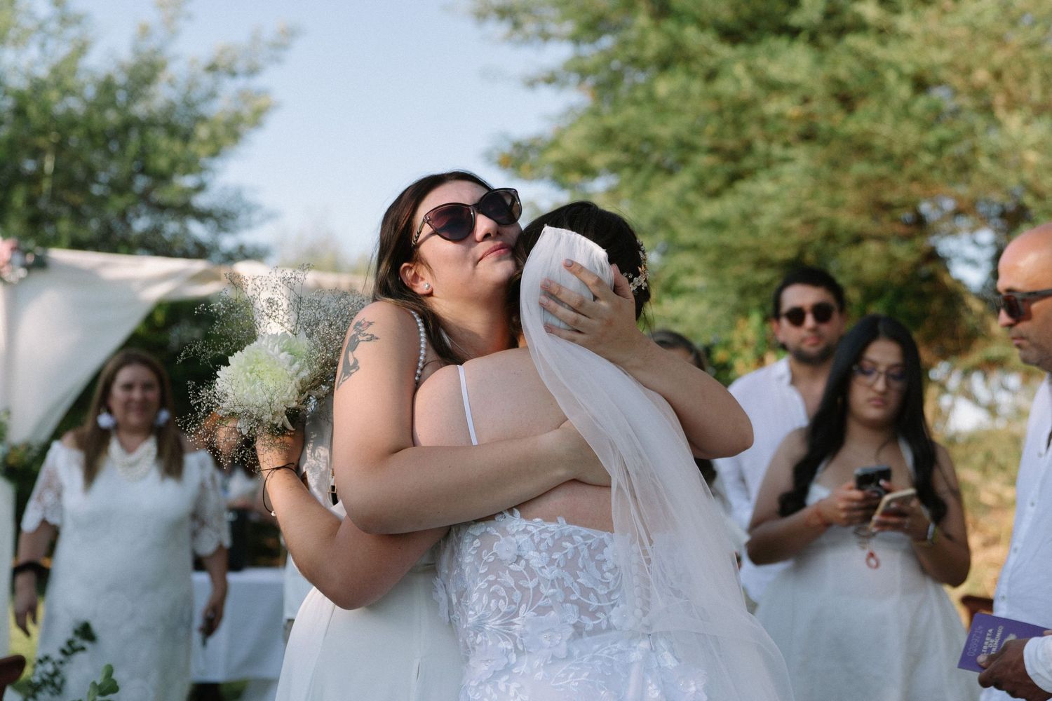 Bride hugging a guest in sunglasses at an outdoor wedding ceremony, holding a white bouquet.