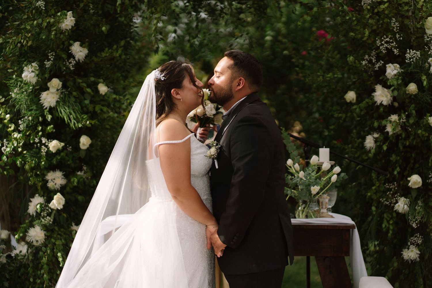 Bride and groom sharing first kiss under white floral arch at outdoor garden wedding.