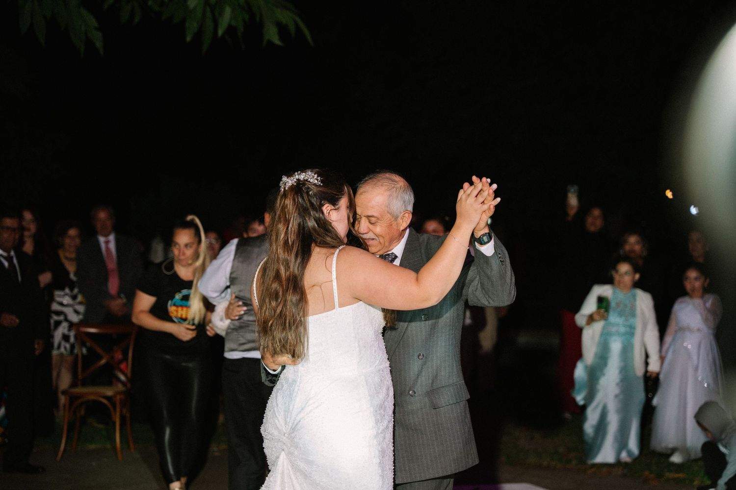 Bride shares emotional father-daughter dance at outdoor evening wedding reception with guests watching.