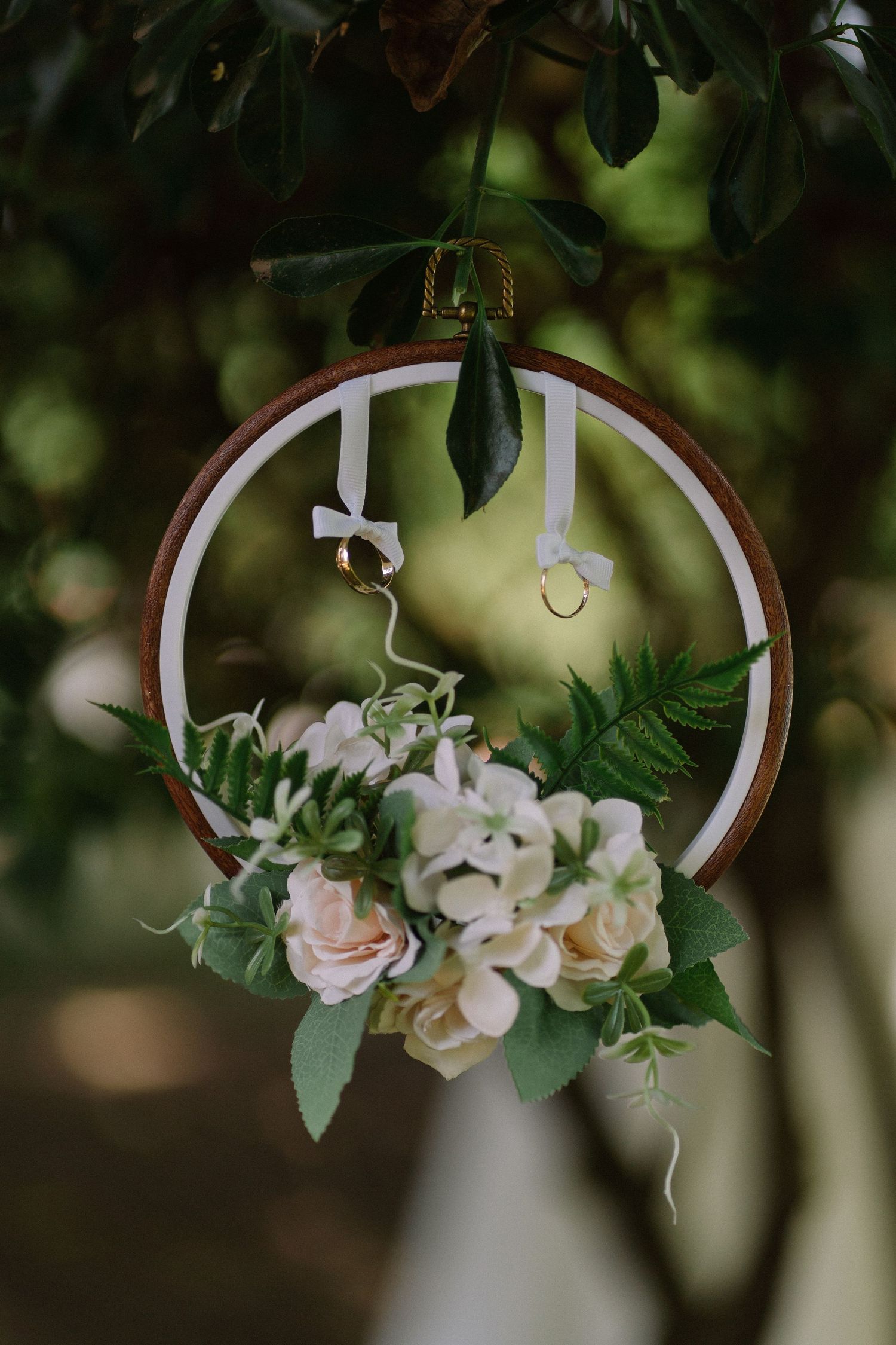Decorative wedding ring hoop with white and pink flowers and gold rings hanging from tree branch.