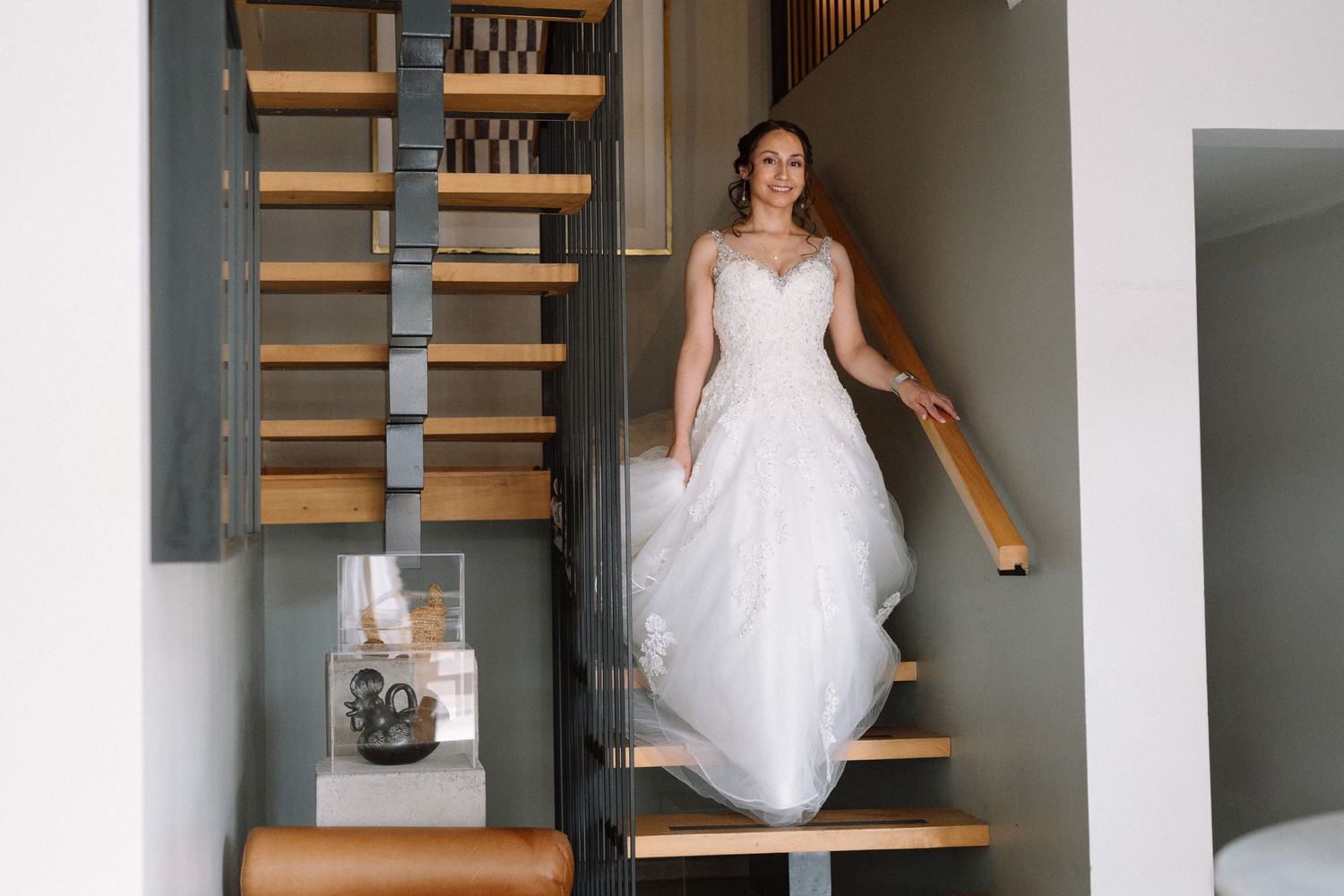 Bride in white wedding gown descending wooden staircase in modern interior.