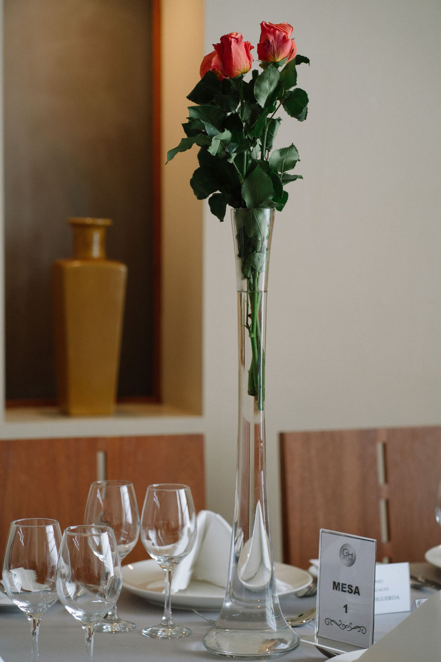 Two red roses in a tall glass vase with wine glasses and place settings on an elegant dining table.