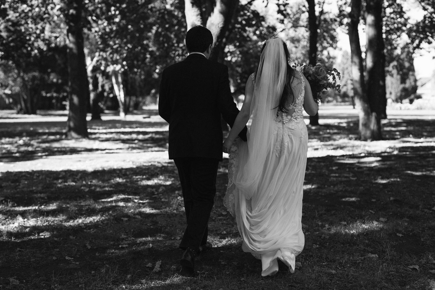 Black and white image of a couple strolling through a park in wedding attire.