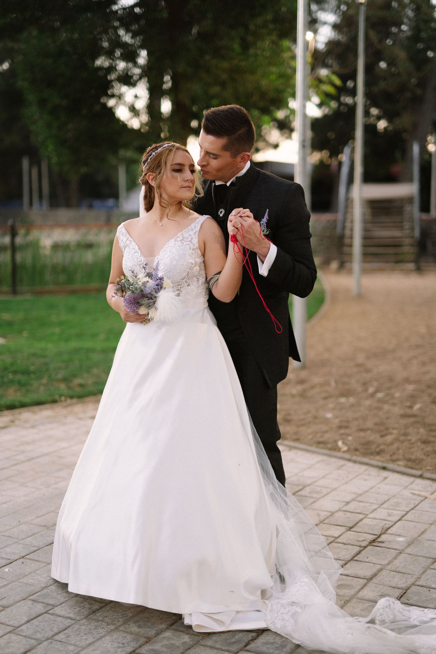 A romantic wedding couple dances with a purple bouquet on a paved path at dusk.
